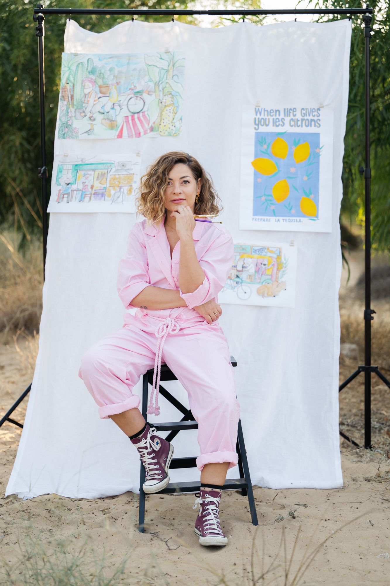A lady in a pink jumpsuit set sits on a black chair against a white backdrop with illustrated artwork displays.