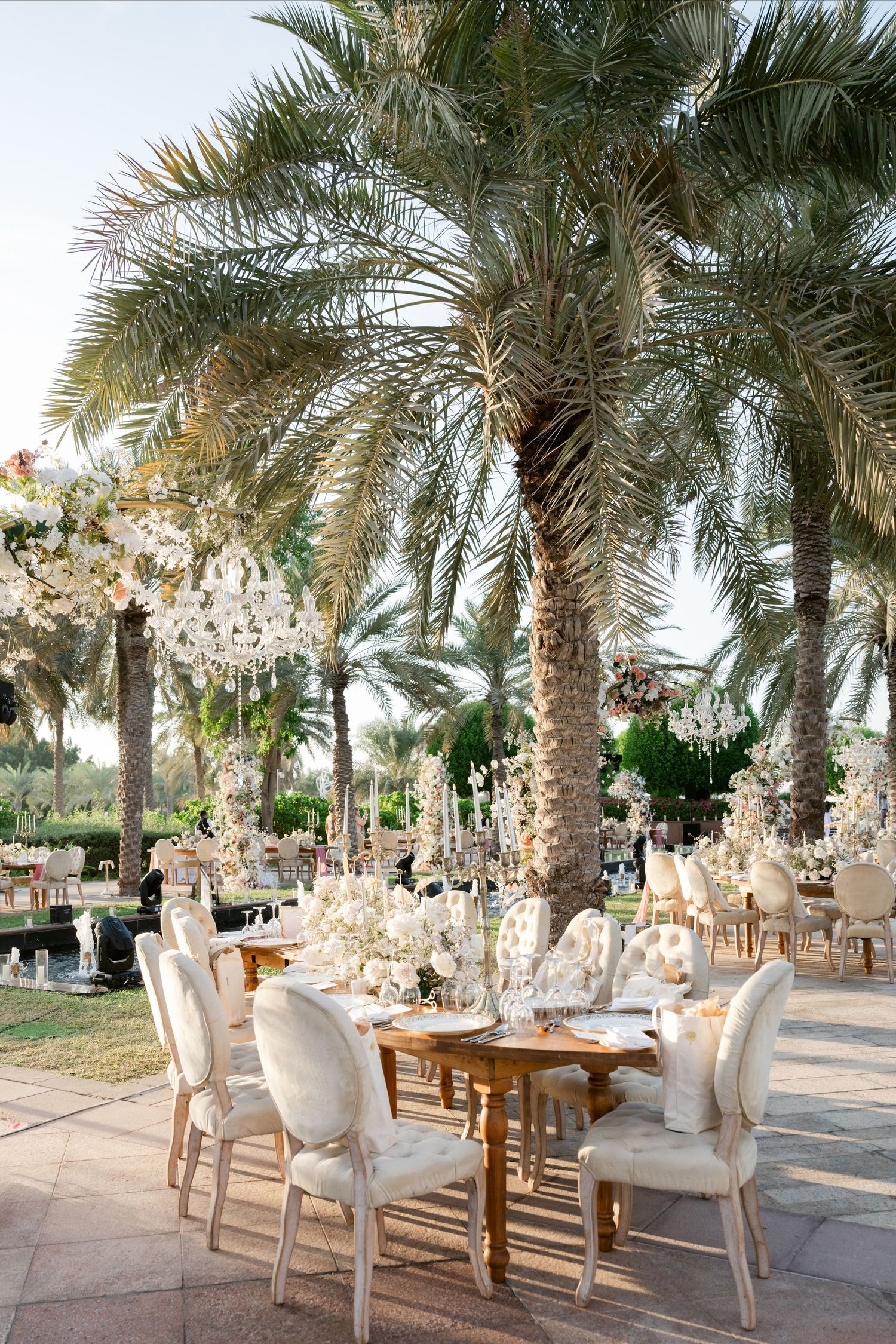 Outdoor wedding reception setup with white chairs and floral arrangements under towering palm trees.