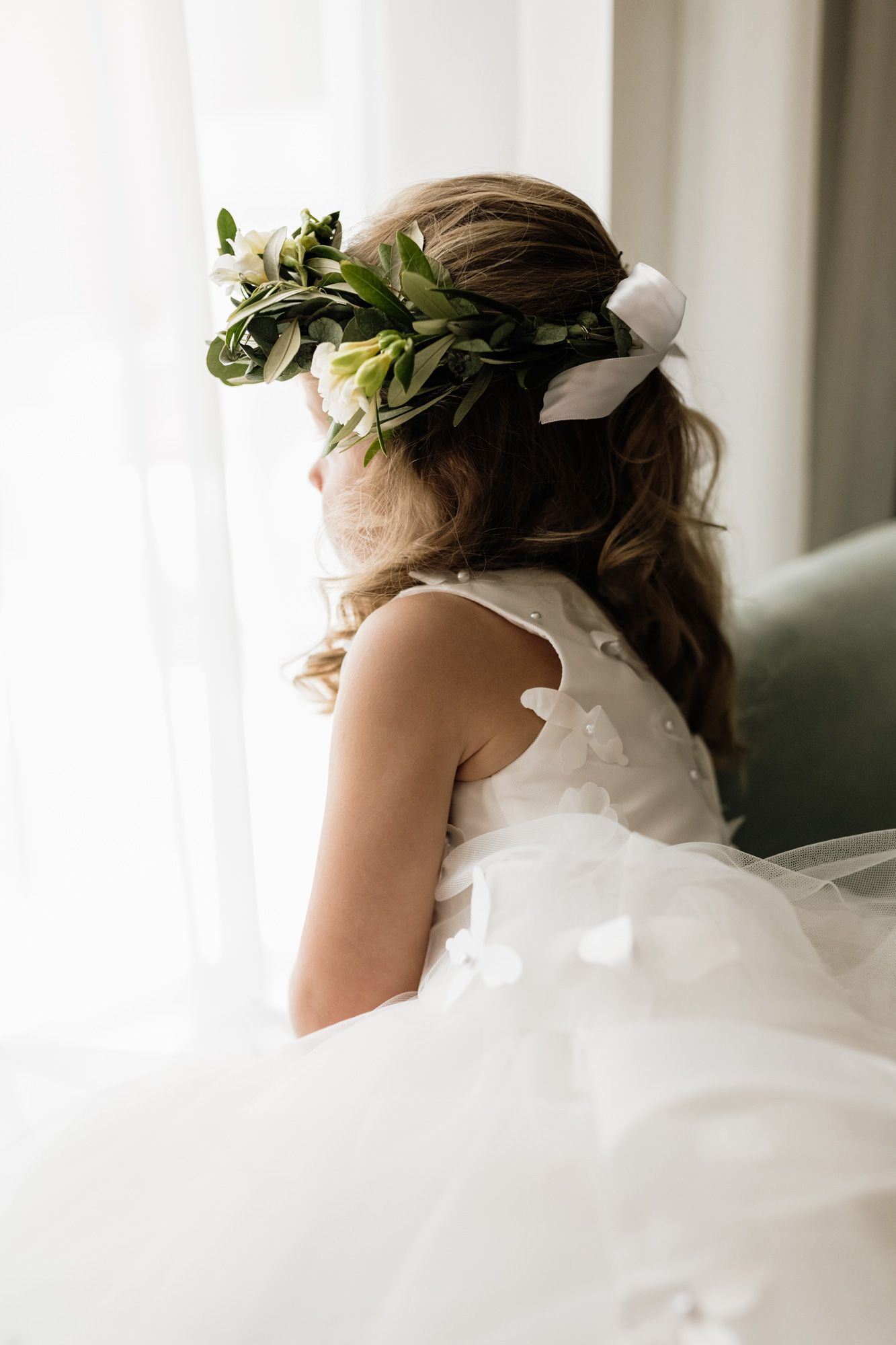 A flower girl wearing a white veil and floral crown looks away from the camera in an elegant side profile portrait.