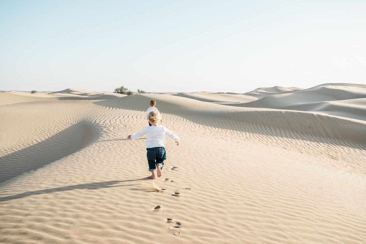 Two boys explore vast desert sand dunes under bright sunlight, leaving footprints in their wake.
