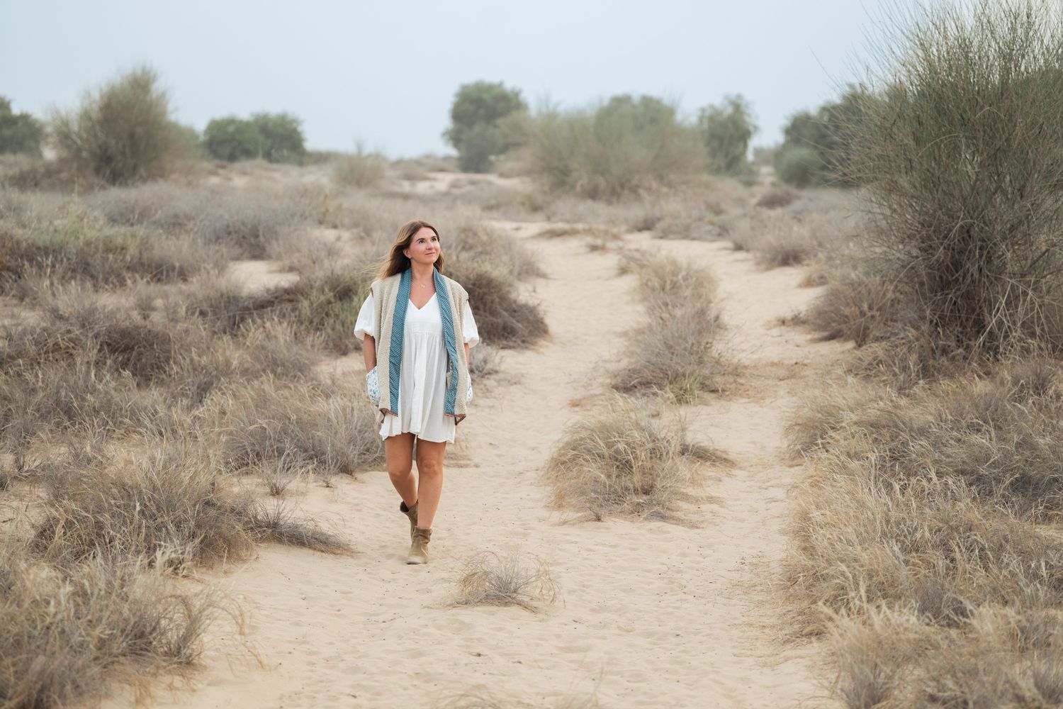 A person in a blue jacket walks with their dog along a winding desert trail surrounded by scrub brush and sand.