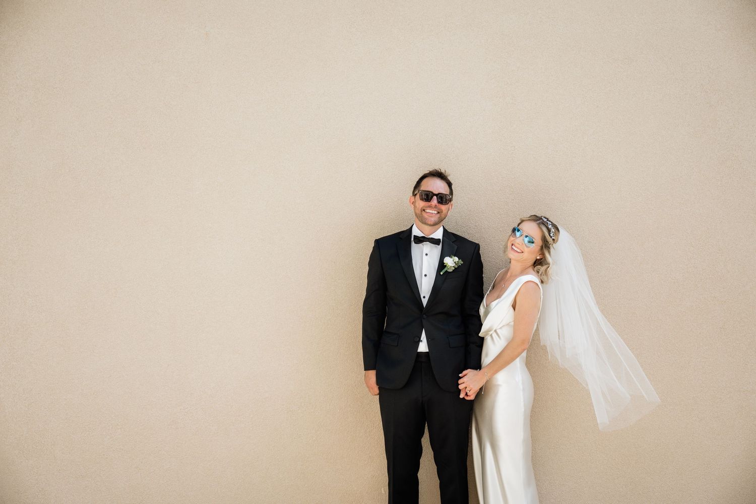 Couple in formal wedding attire posing against beige wall.
