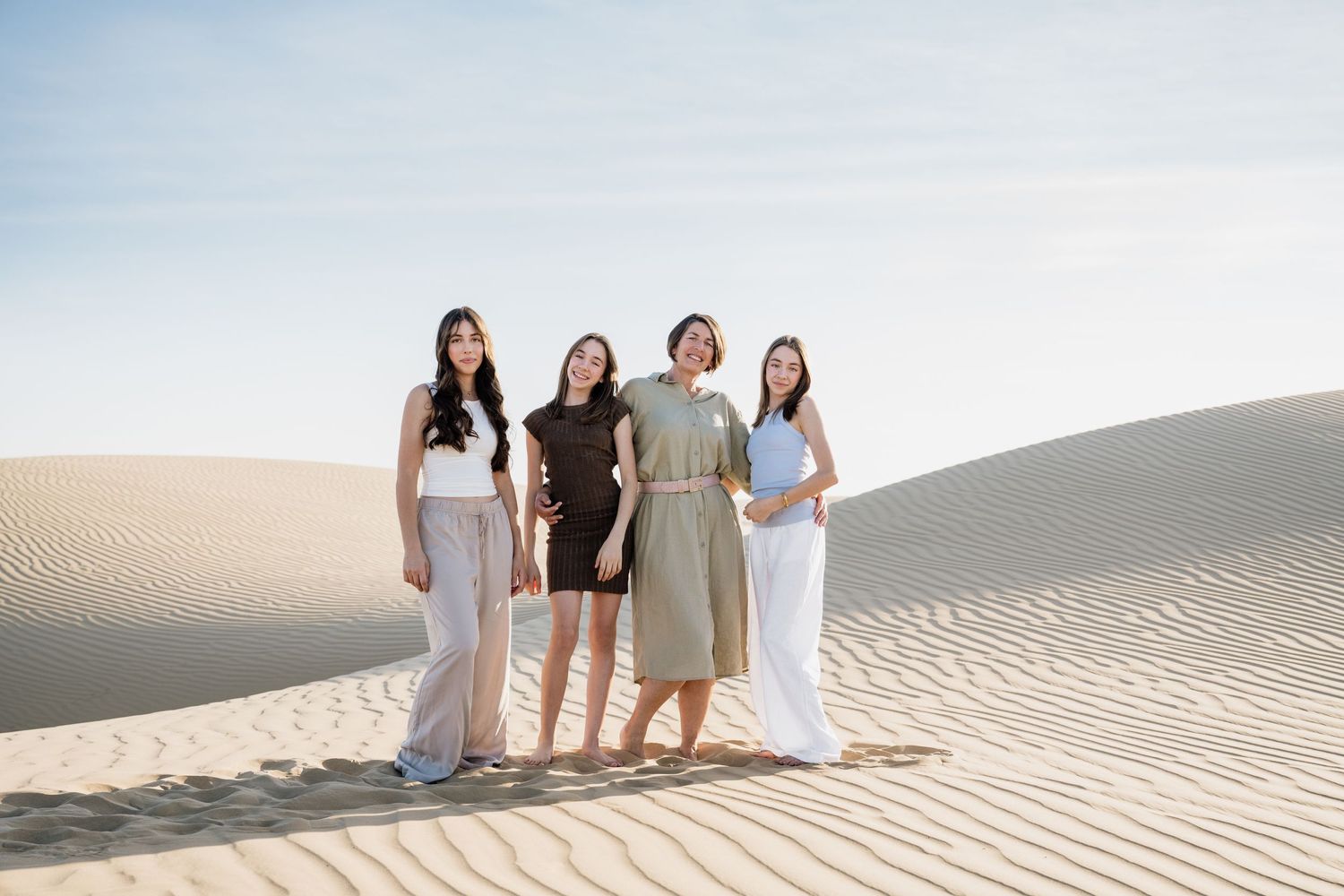 Friends in light colored outfits pose together on rippling sand dunes under bright sky.