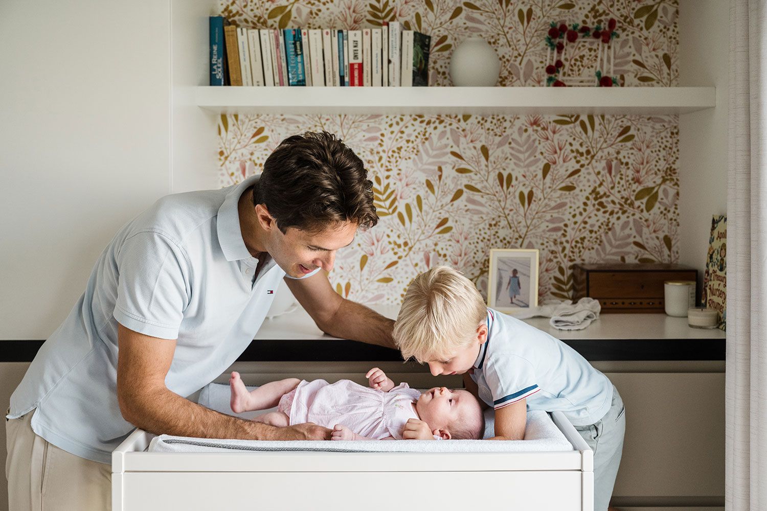 Father and son lovingly tend to a baby on a white changing table with floral wallpaper and shelving in the background.