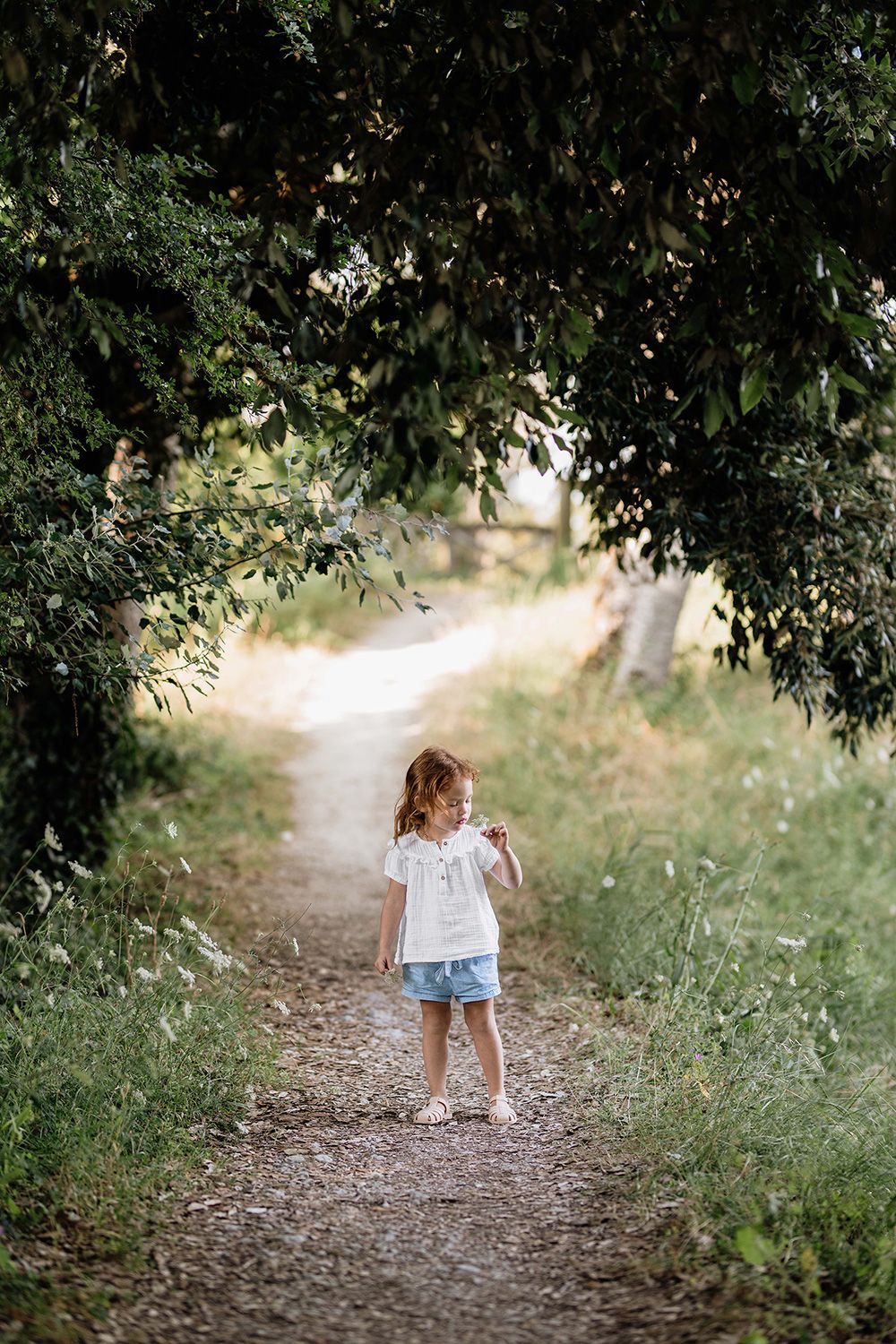 A small child in a white dress walks down a shaded dirt path through trees and greenery.