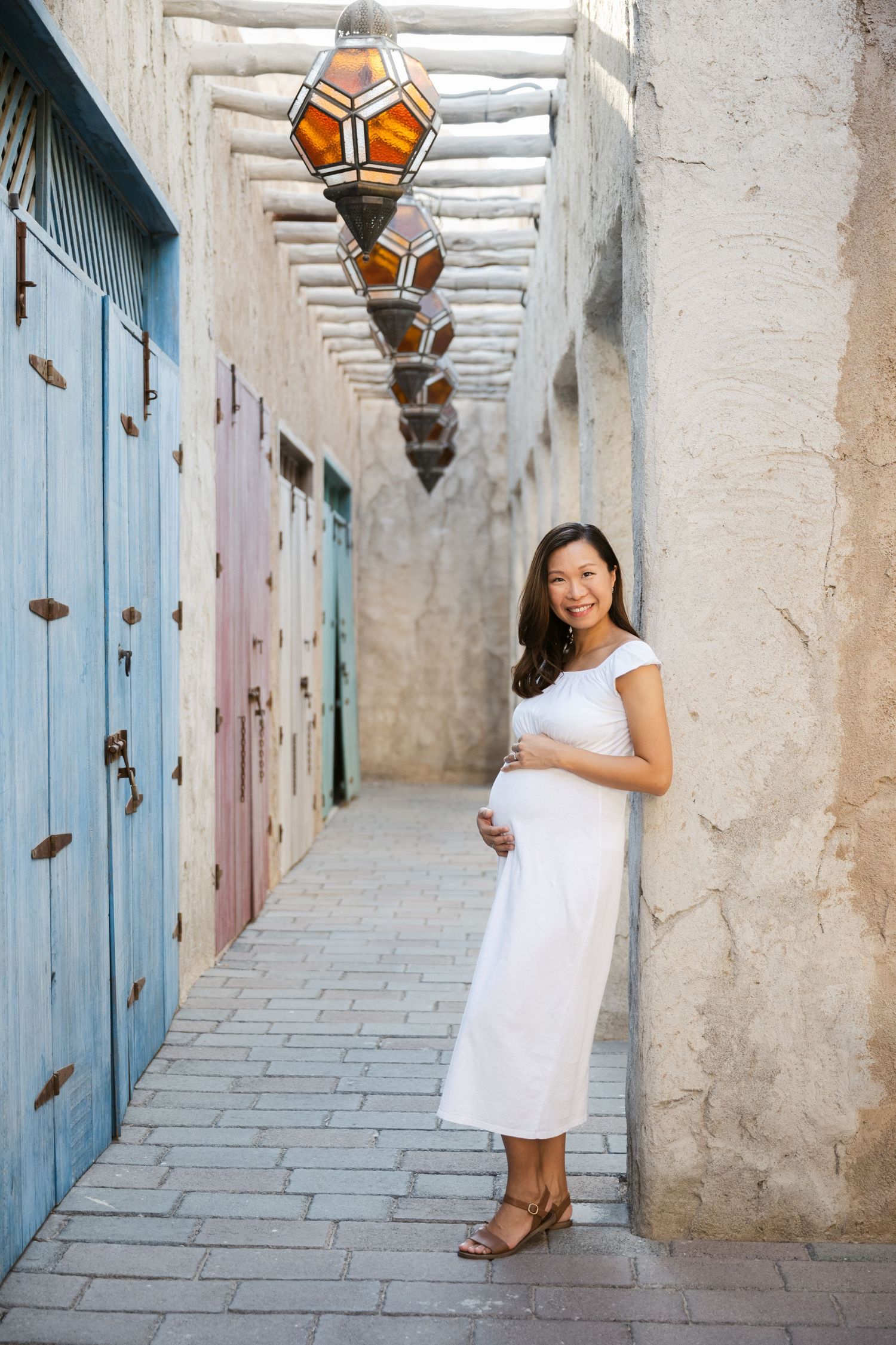 Woman in white dress poses against rustic wall with blue doors and decorative lanterns in Mediterranean setting.