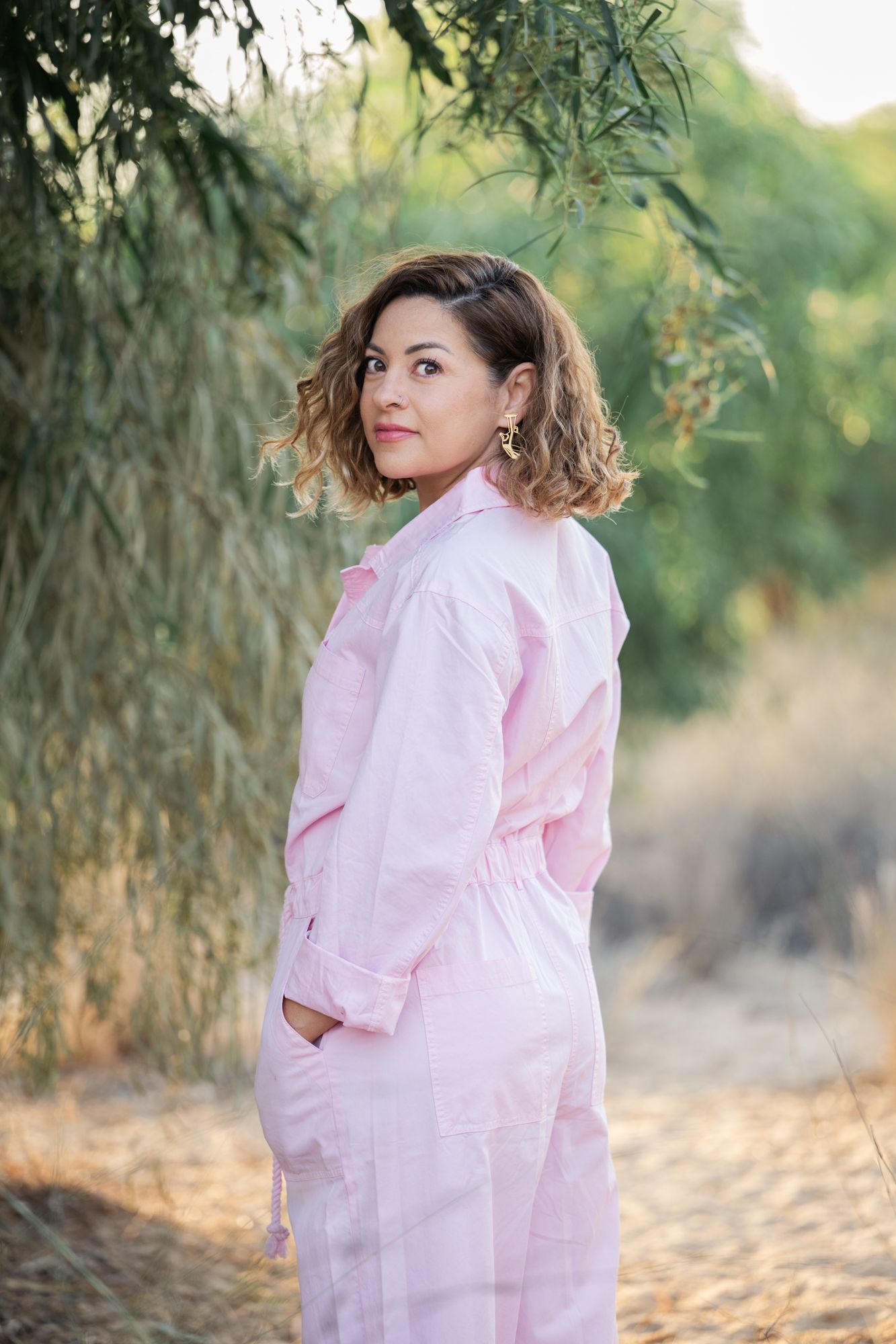 A person in a soft pink dress poses gracefully outdoors against a natural backdrop of large trees and sunlight.