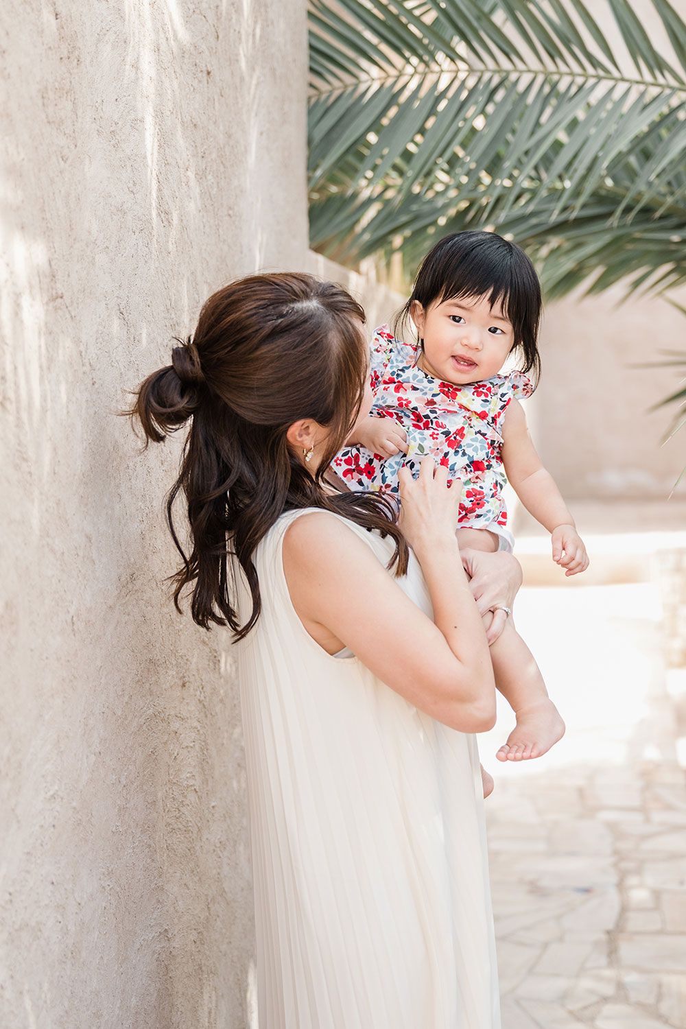 A child in a floral dress peeks playfully around in her mother's arms in Old Dubai