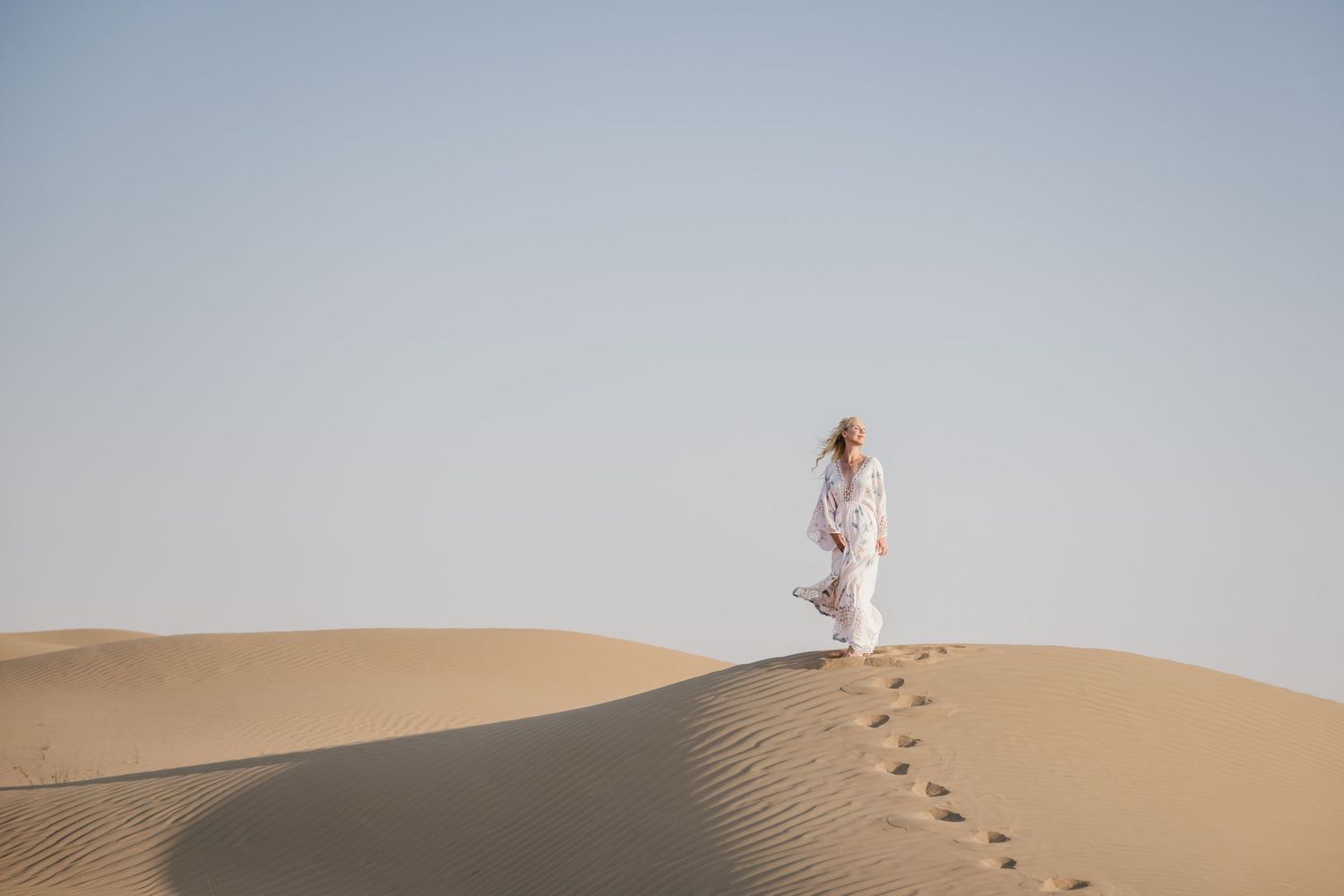 Person in white clothing walking across sandy desert dunes at sunset creating long shadows.