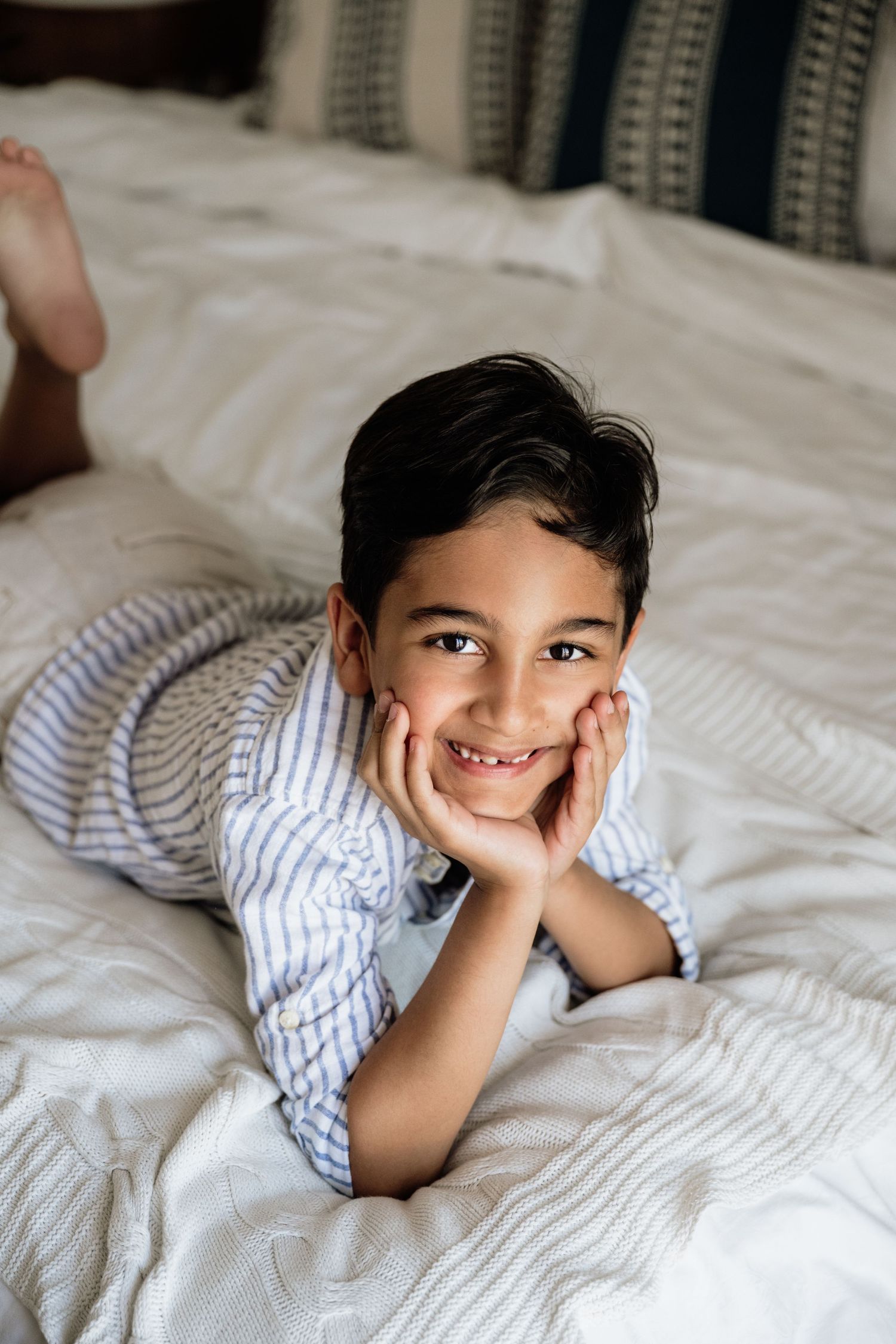 Child laying on white bedding with joyful expression and hand under chin.