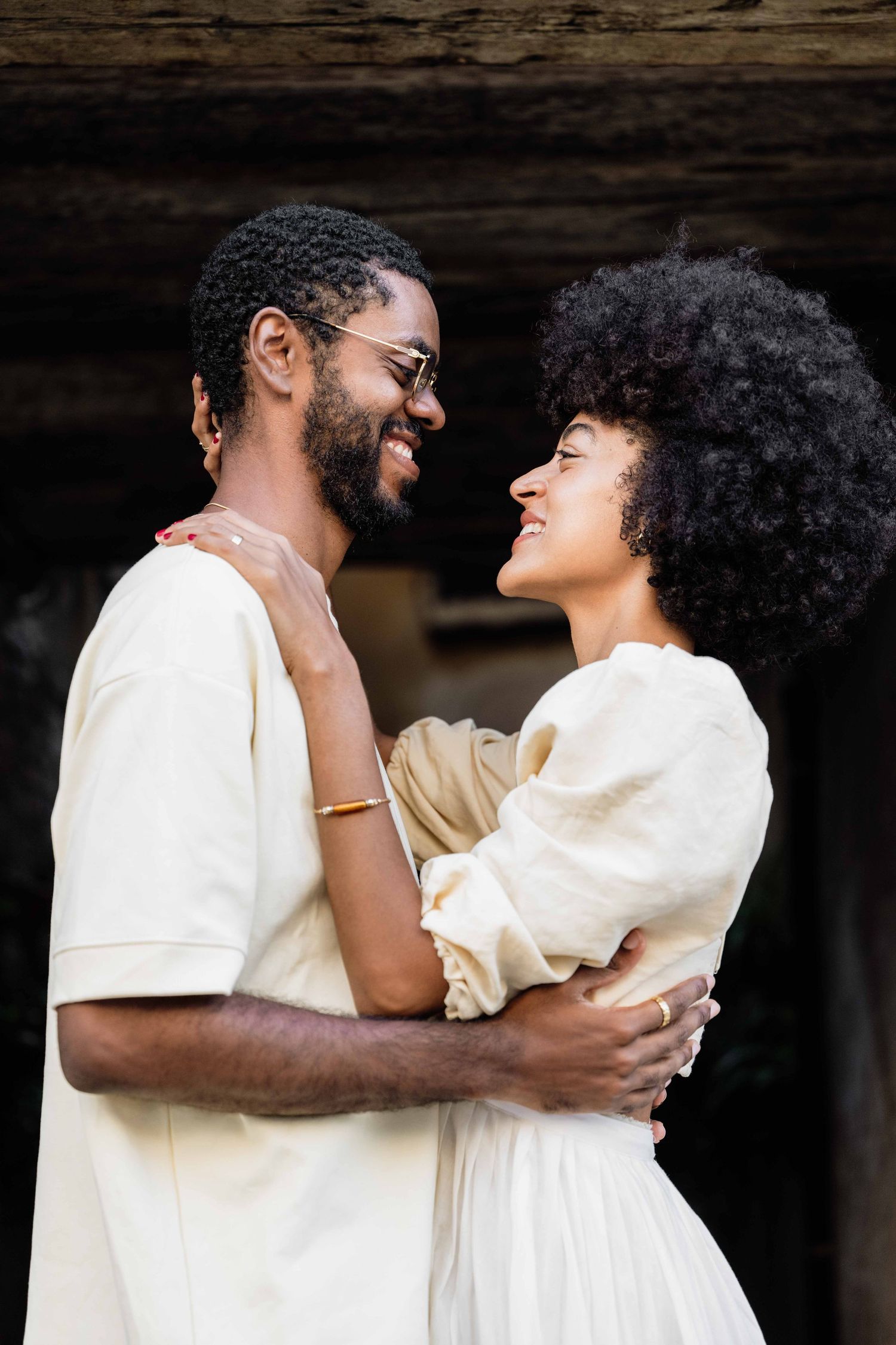 Joyful couple in white clothing sharing laughs and embraces in dim lighting.