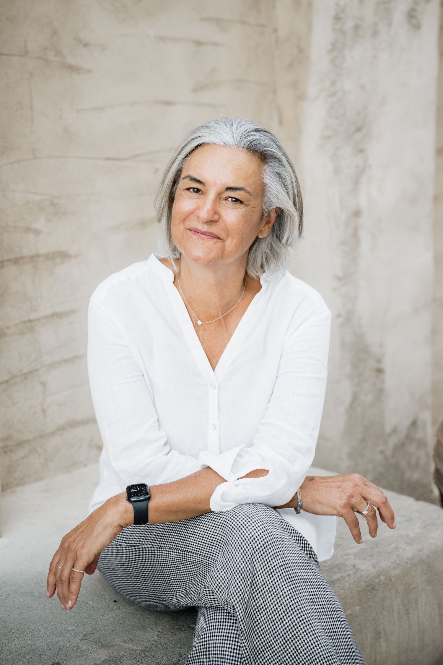 Woman in white shirt and checkered pants relaxing against concrete wall.