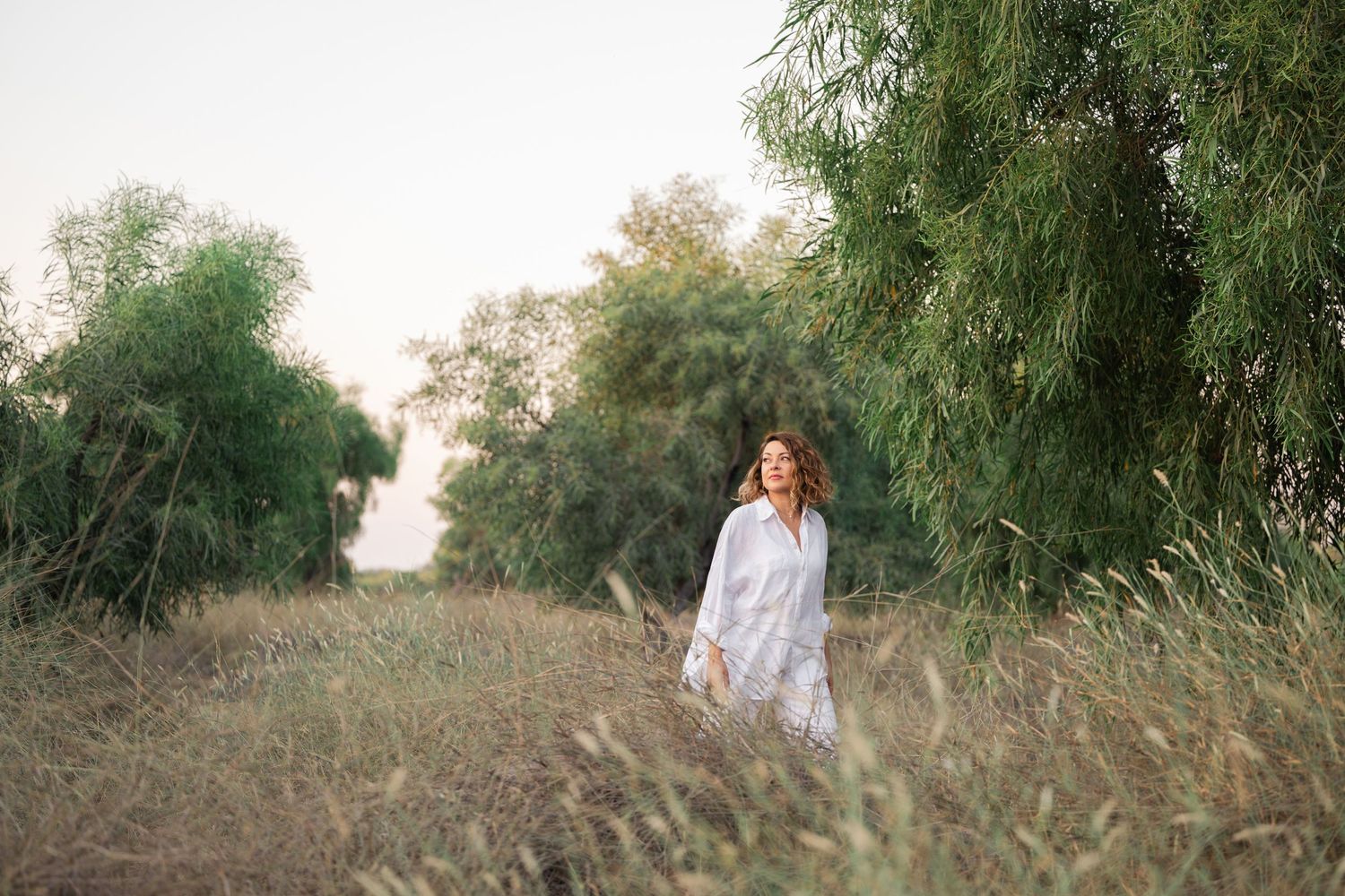 A couple walks together through a serene natural setting with tall grass and trees during golden hour.