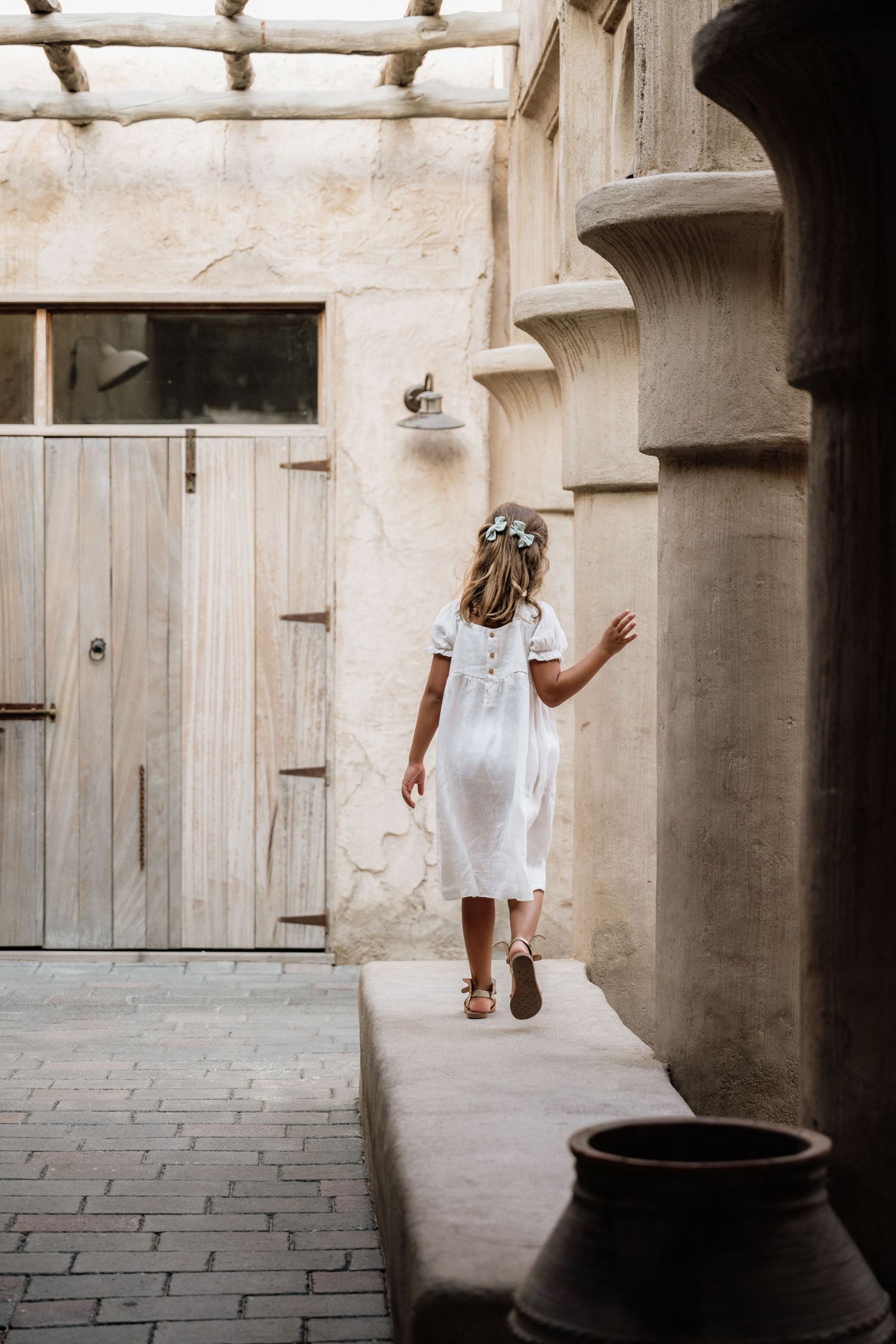 A figure in a flowing white dress and floral crown skips through a rustic stone archway near a wooden door.