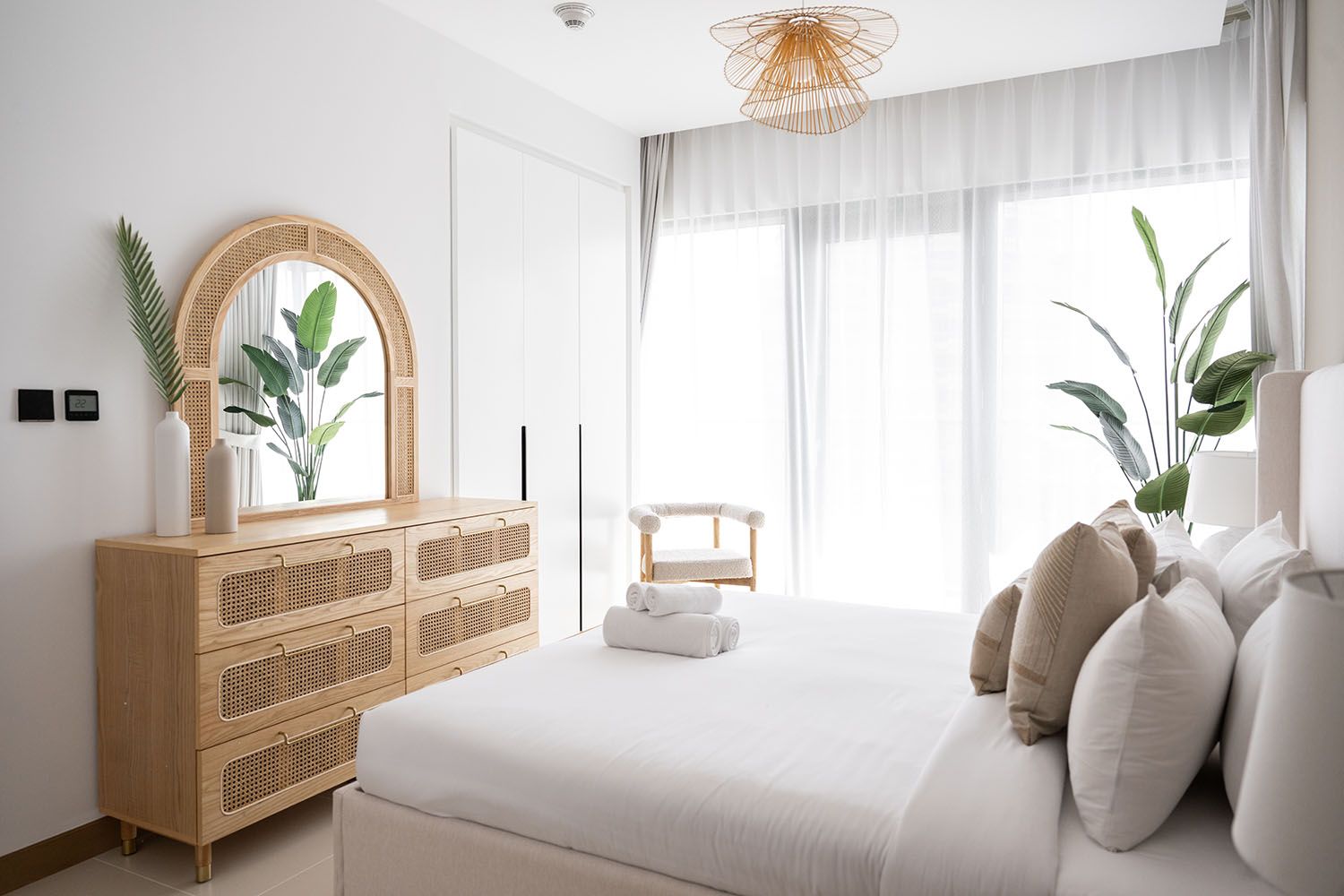 Bright coastal bedroom with rattan furniture, white bedding, and natural light streaming through sheer curtains.