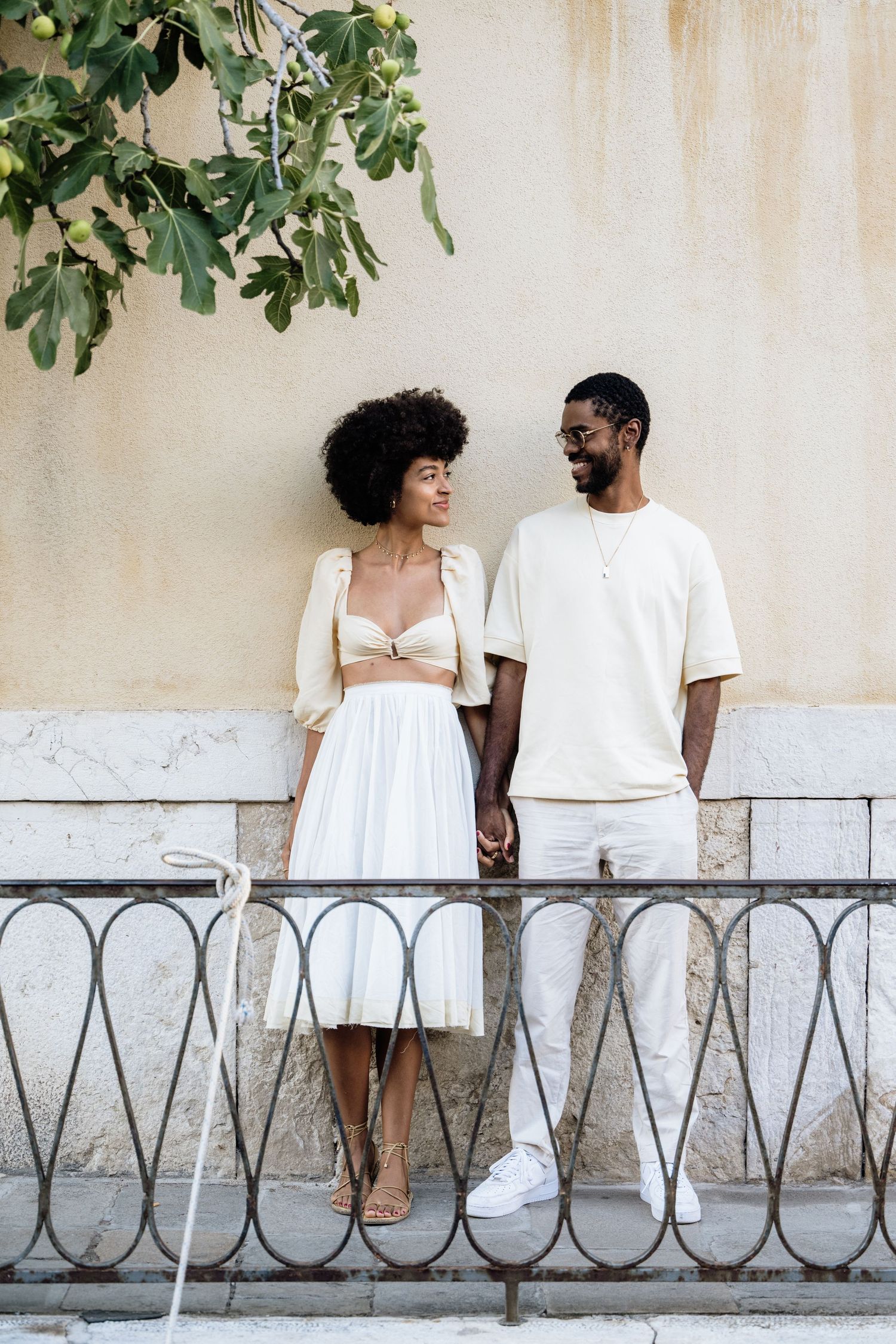 Romantic couple in white outfits sitting together against a light colored wall with decorative railing.