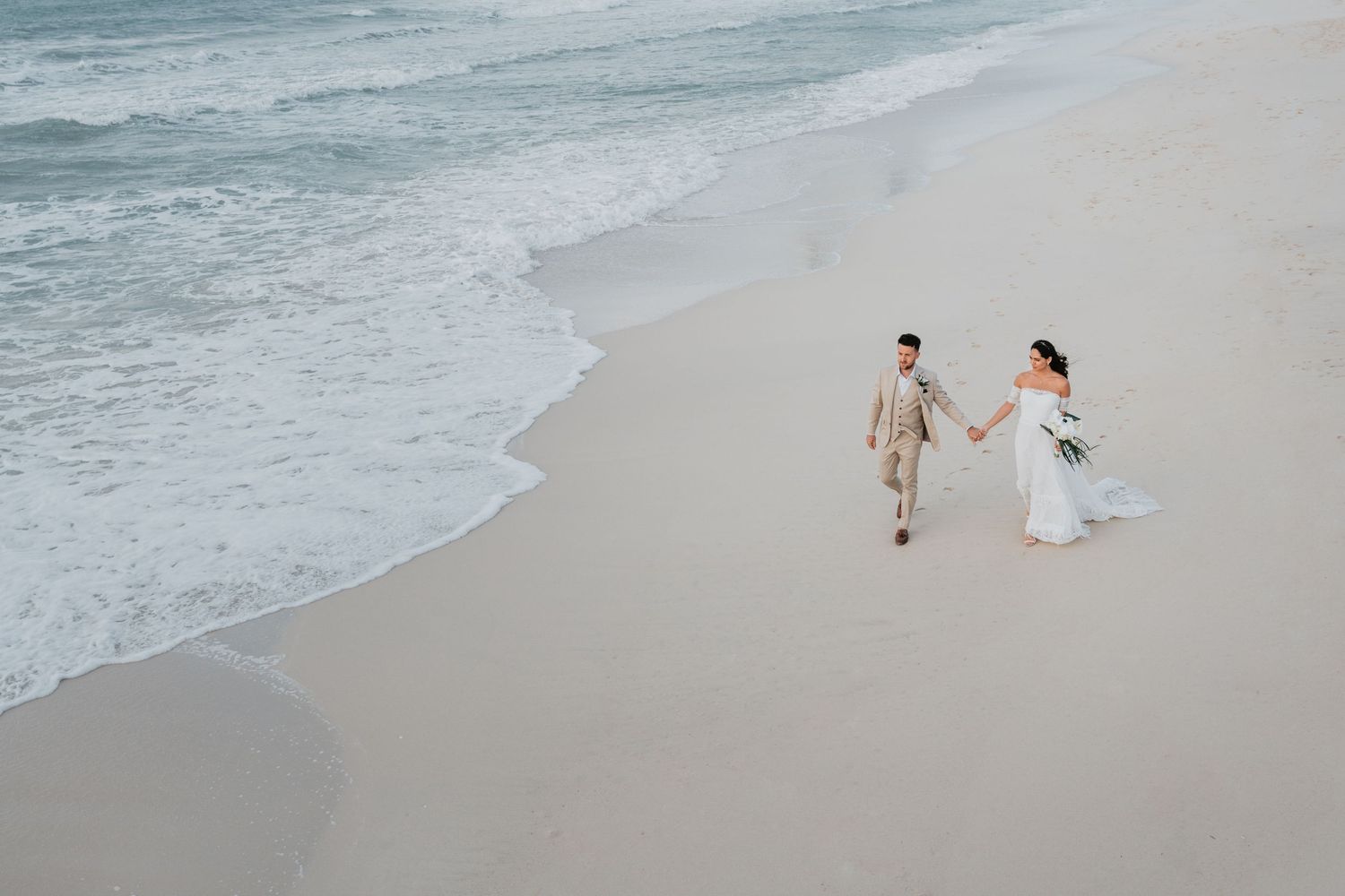 A couple in wedding attire walks along a serene beach shoreline with ocean waves rolling in.