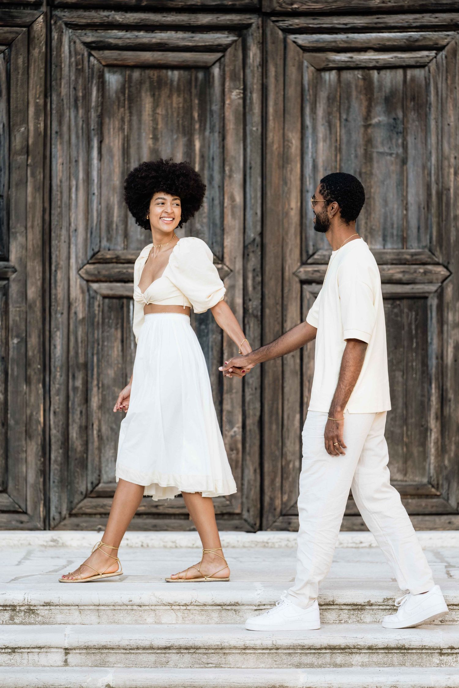 A stylish couple in matching white outfits poses on stone steps near vintage wooden doors.
