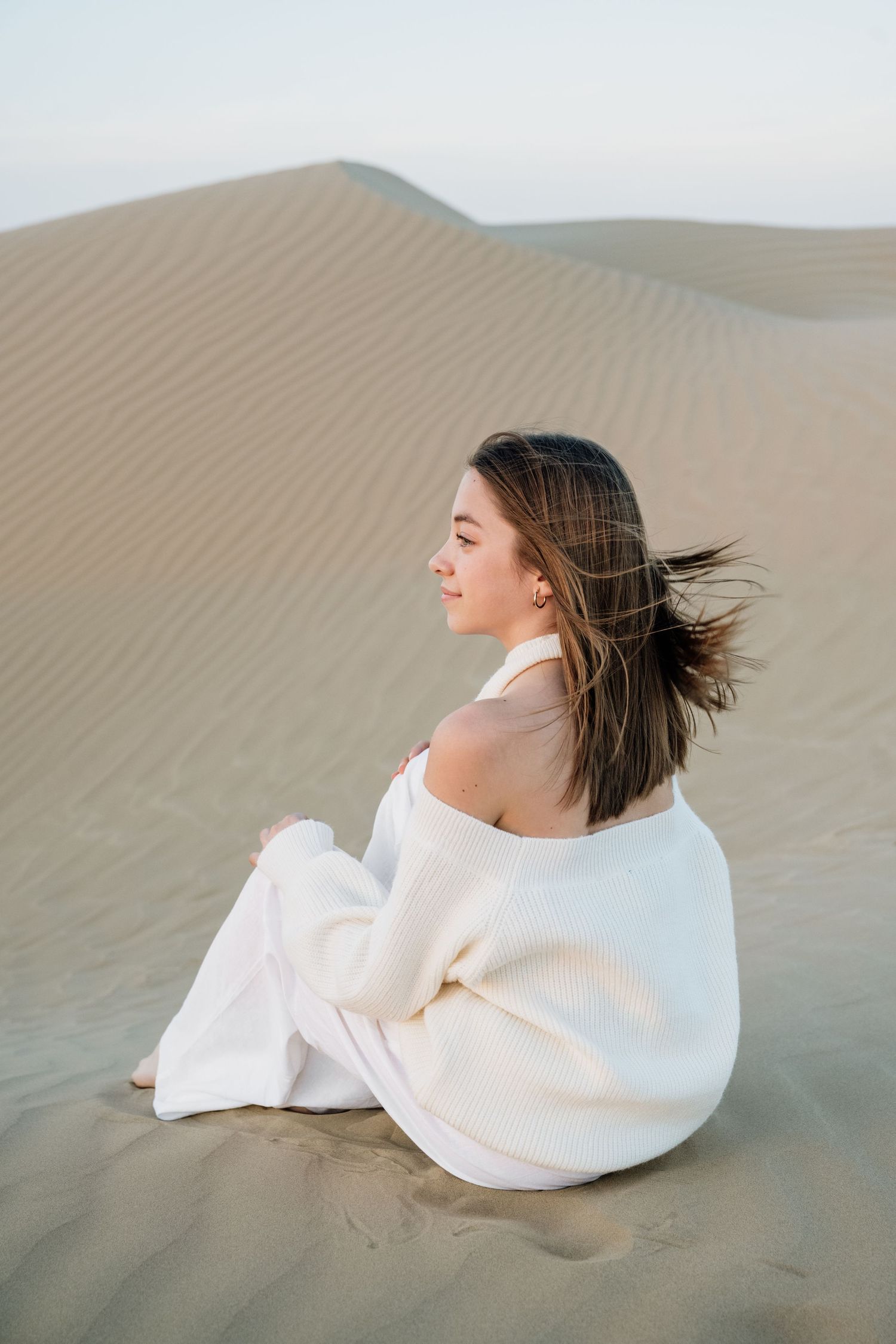 Person in flowing white dress lying gracefully on sand dunes during a desert photoshoot.