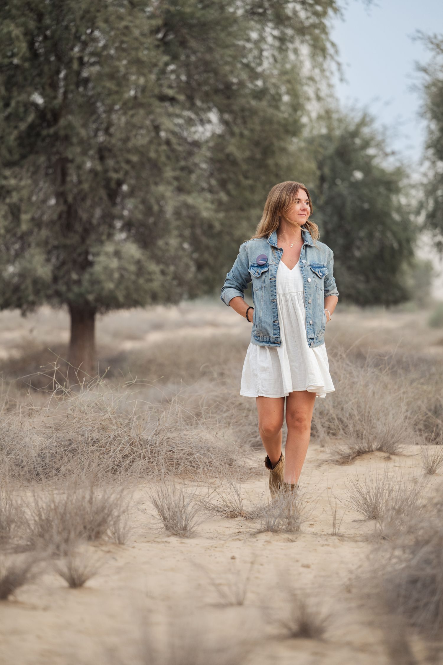Woman in white dress and denim jacket walks through desert landscape with scattered trees in background.