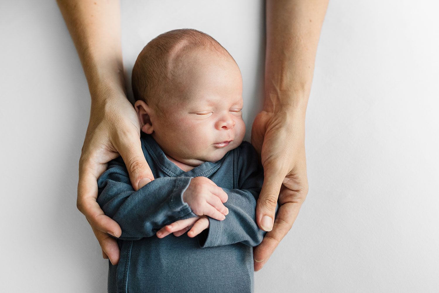 A sleeping newborn baby in blue clothing is gently cradled in adult hands.