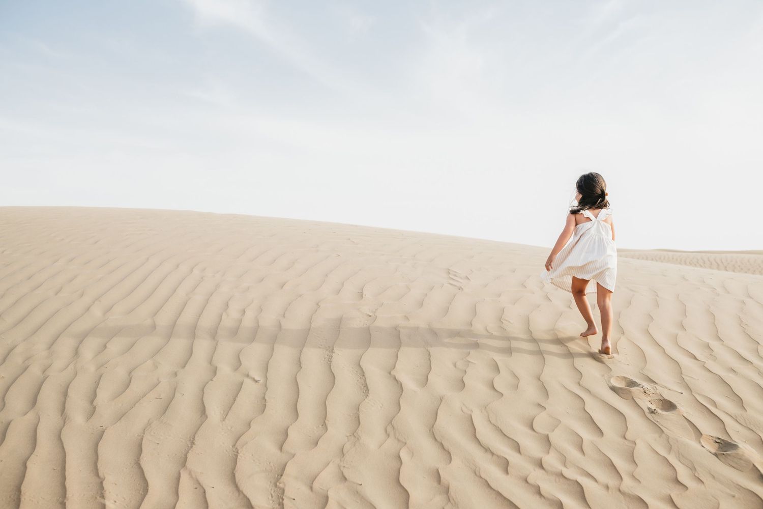 A person in a white dress walks across rippling sand dunes in a desert landscape.