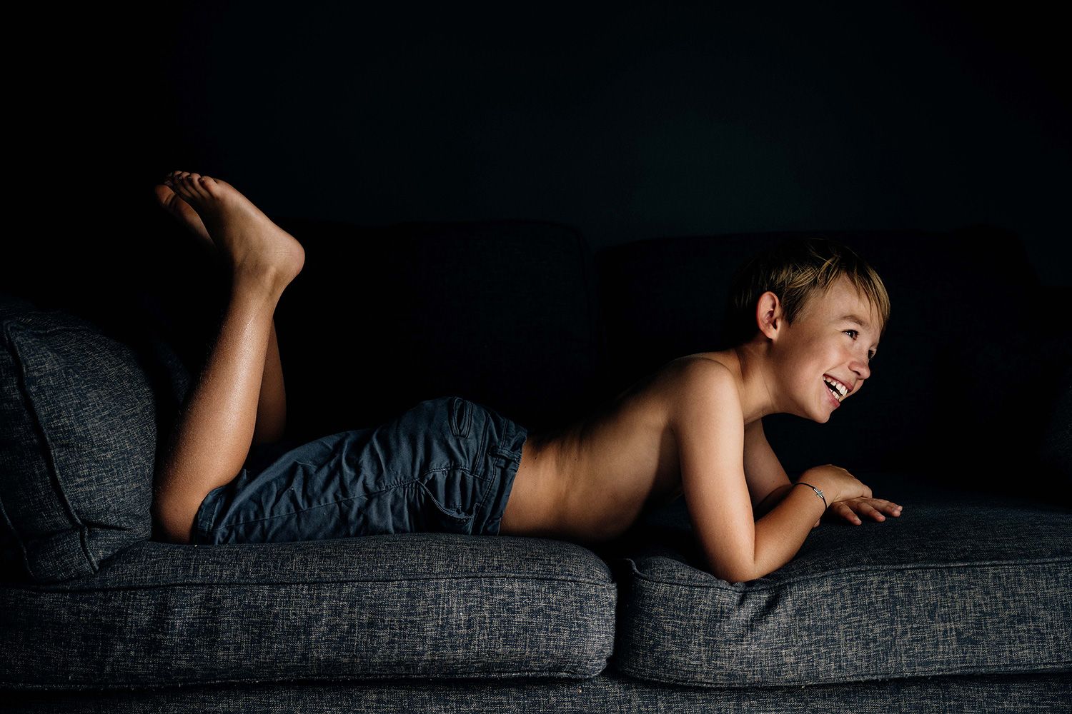A person lies on a dark couch in a dramatic boudoir pose with moody lighting.
