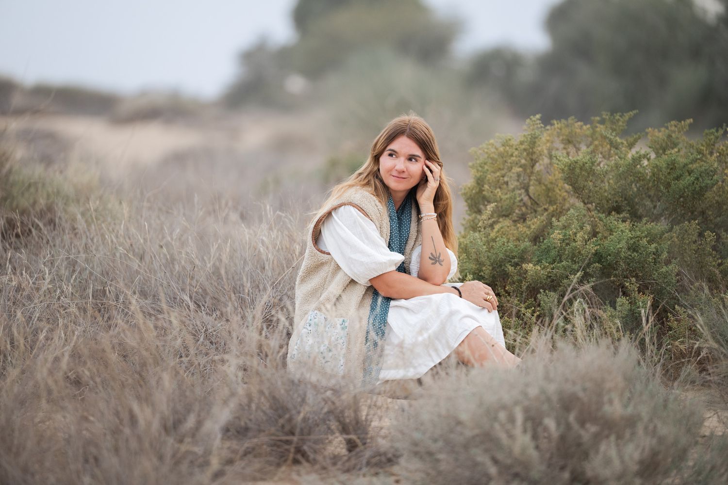 A person in a white dress sits gracefully among tall grass and wildflowers during a dreamy outdoor photoshoot.