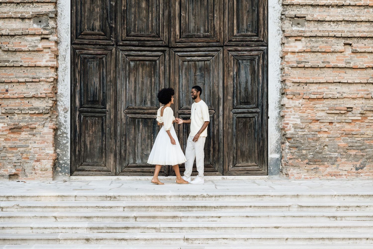 Couple posing against rustic wooden doors and brick wall in romantic photoshoot.