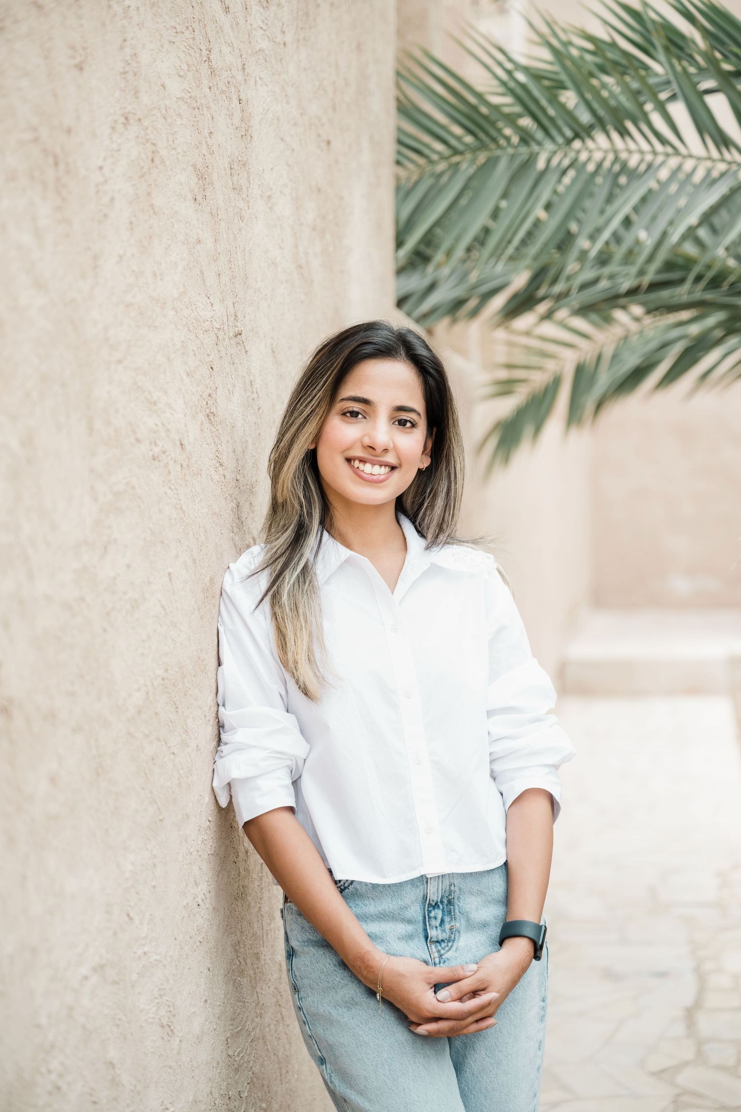 Woman in casual white shirt and jeans poses against beige wall with palm fronds in background.