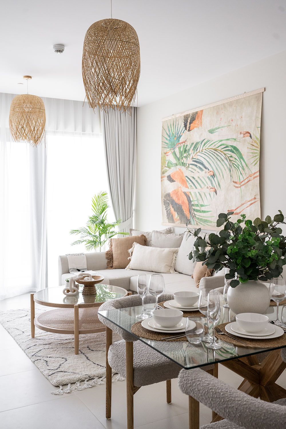 Coastal dining area with glass table, woven pendant light, and tropical artwork on white walls.