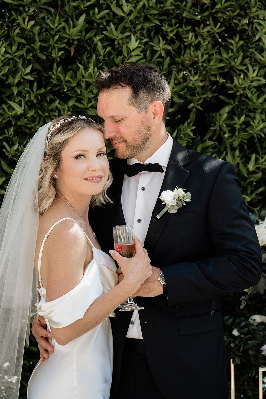 Newlyweds sharing intimate moment against green hedge backdrop during reception.