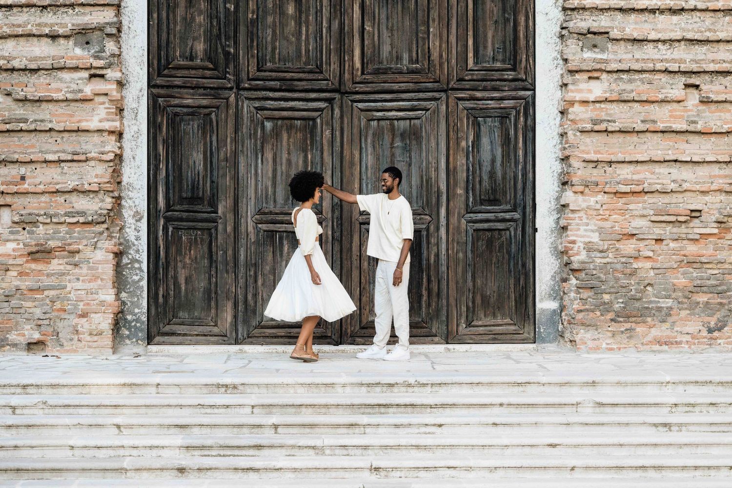 A couple in white outfits dances on stone steps in front of a weathered wooden door.