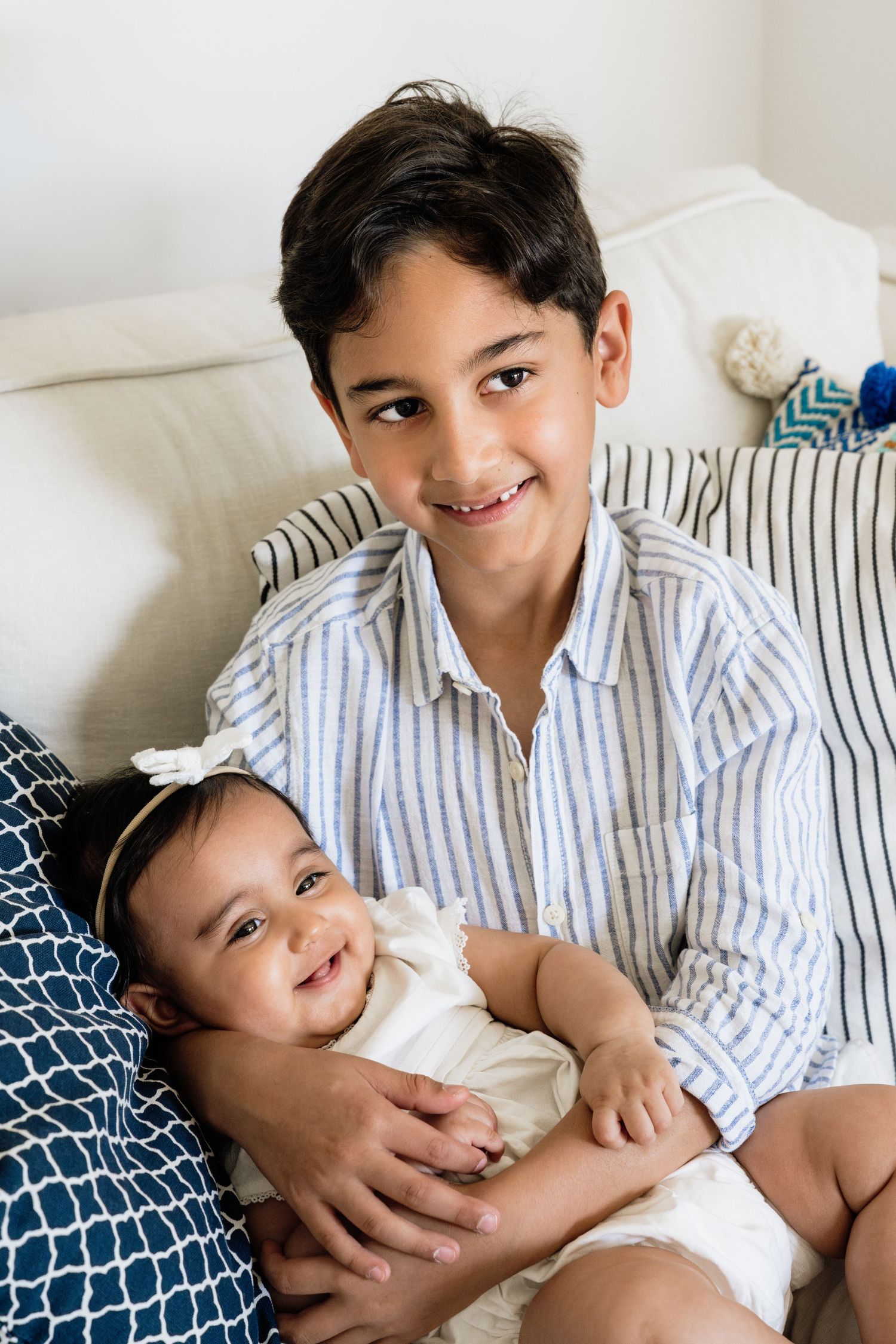 Two siblings sharing tender moment on white couch with decorative pillows.