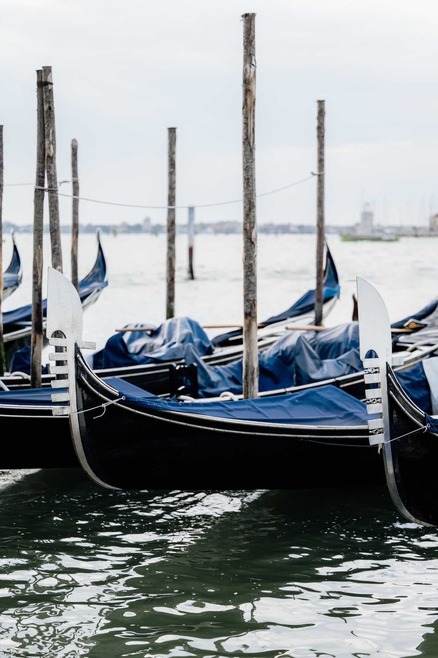 Row of covered gondolas moored at wooden posts in Venice's lagoon with city skyline in the distance.