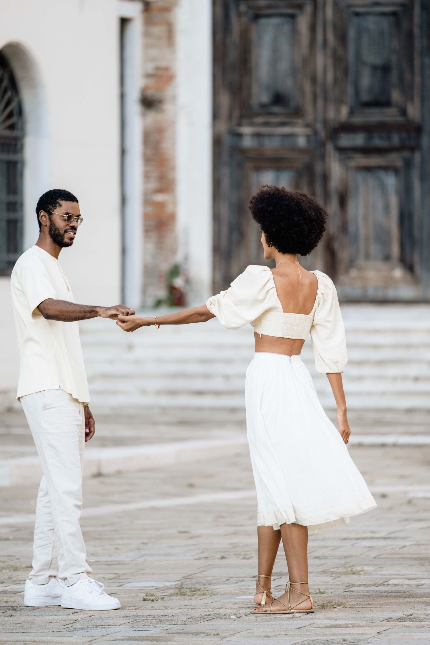 A couple in white outfits dancing playfully in front of a historic building.