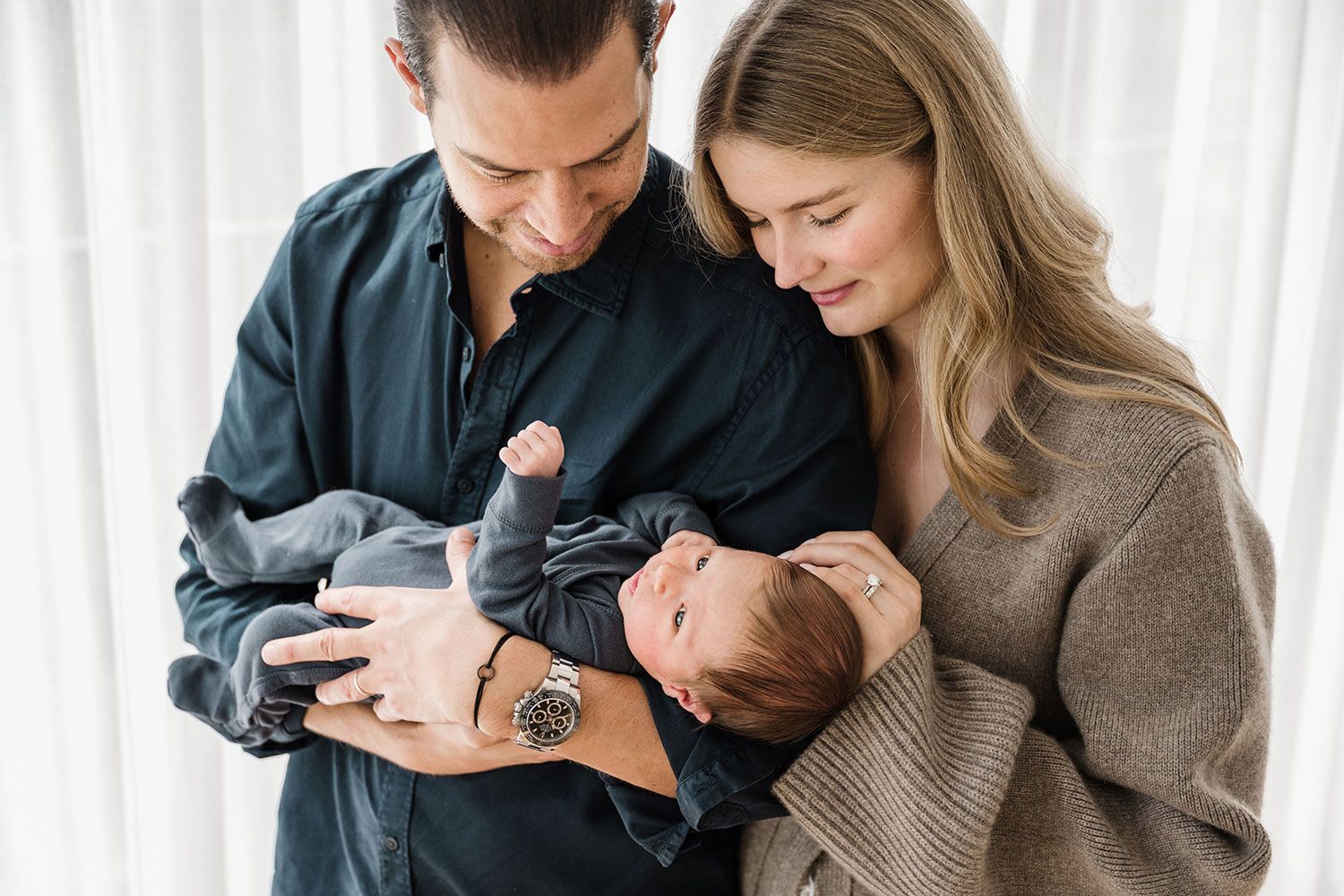 Couple in casual attire stand close together in front of a window holding and looking at their newborn baby