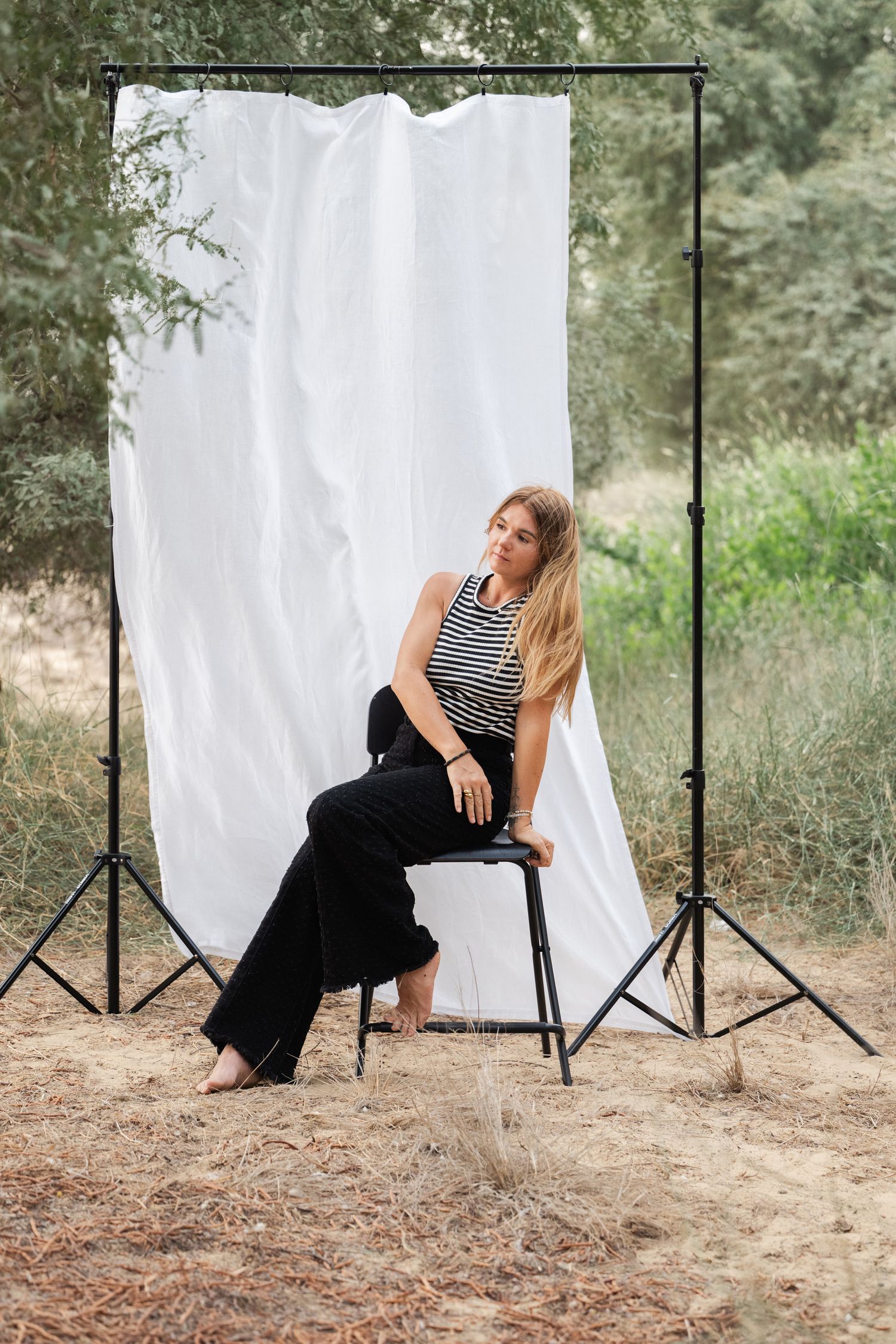 A model in a striped top and black pants poses against a white backdrop in an outdoor forest setting.
