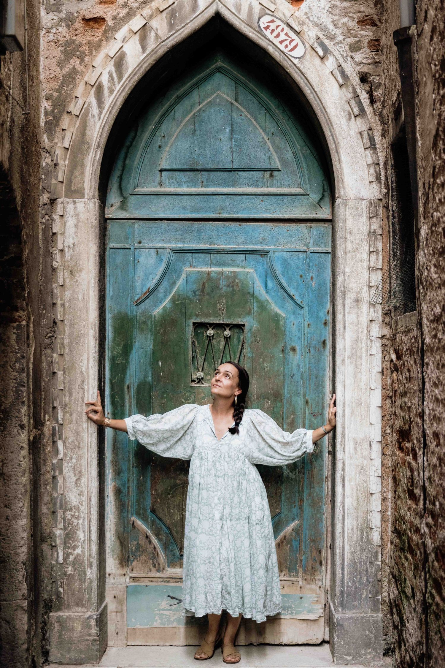 Figure in flowing white dress poses dramatically against arched turquoise doorway in Venice.