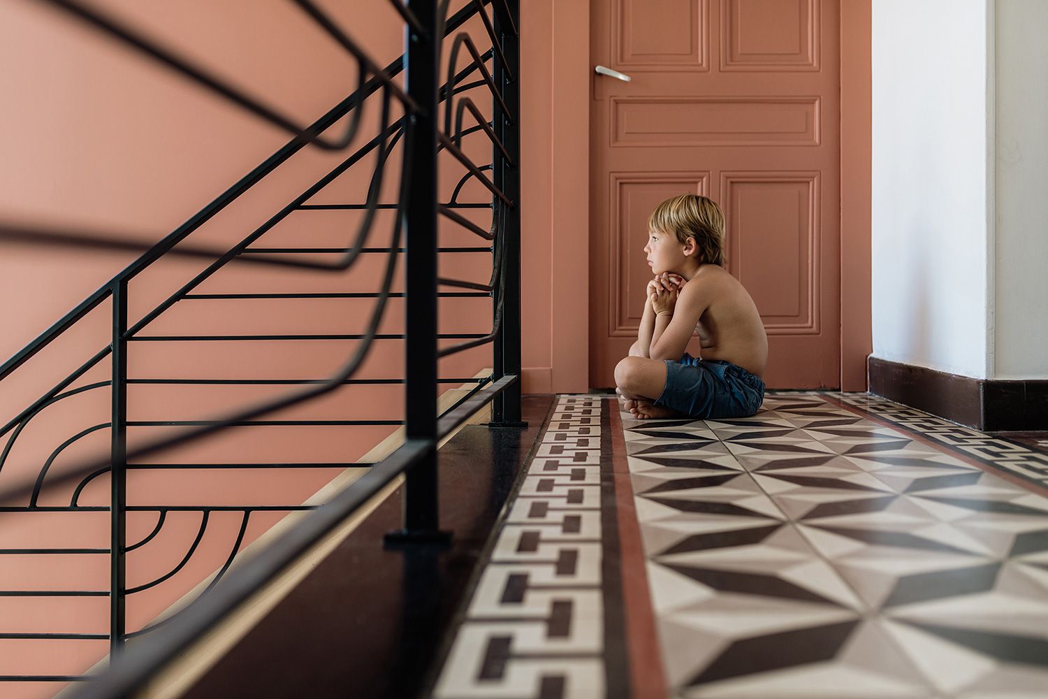 Someone sits alone in a hallway with decorative tiled flooring and a black metal staircase railing.