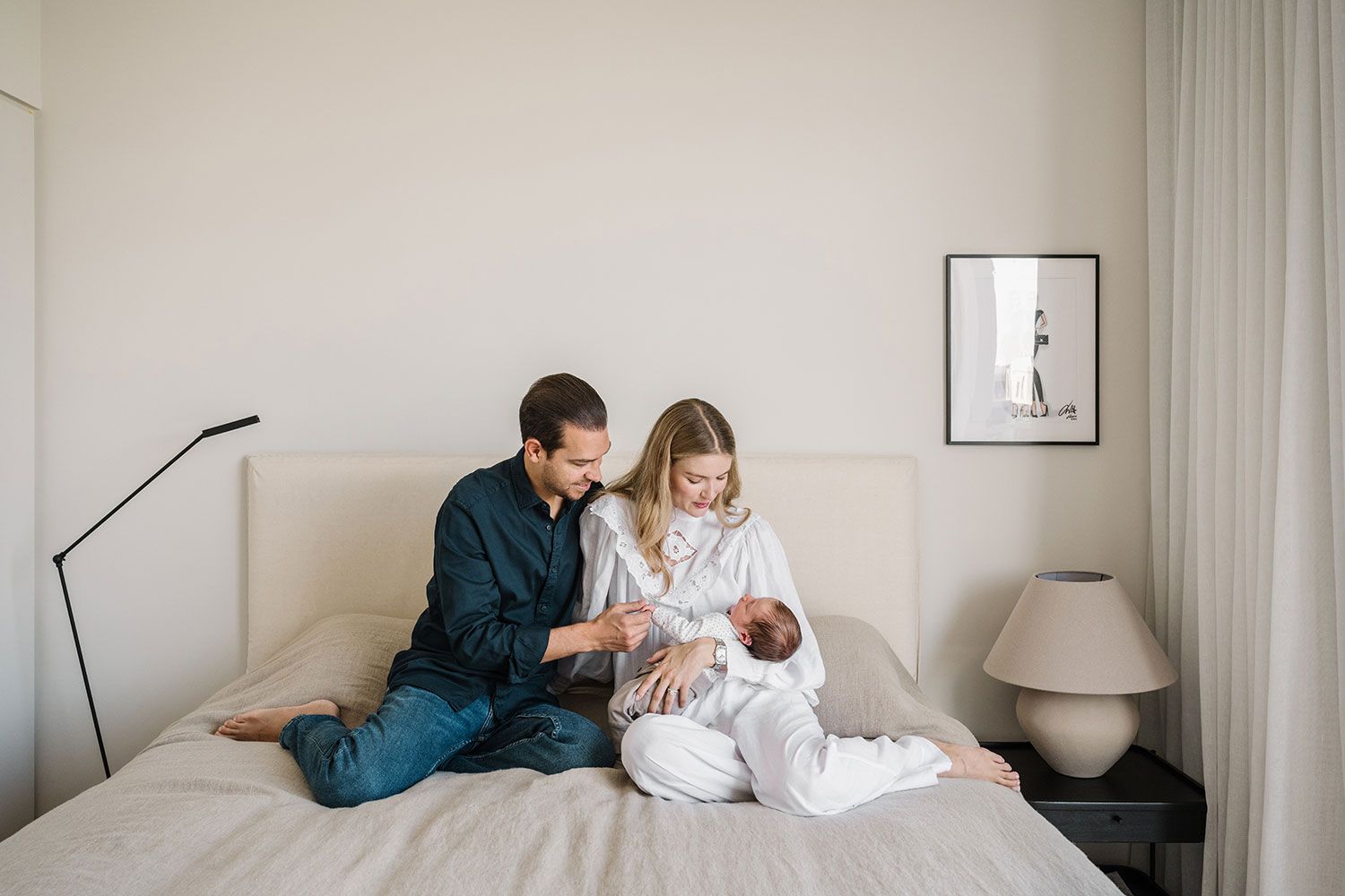 Parents and newborn baby share tender moments in a minimalist white bedroom setting.