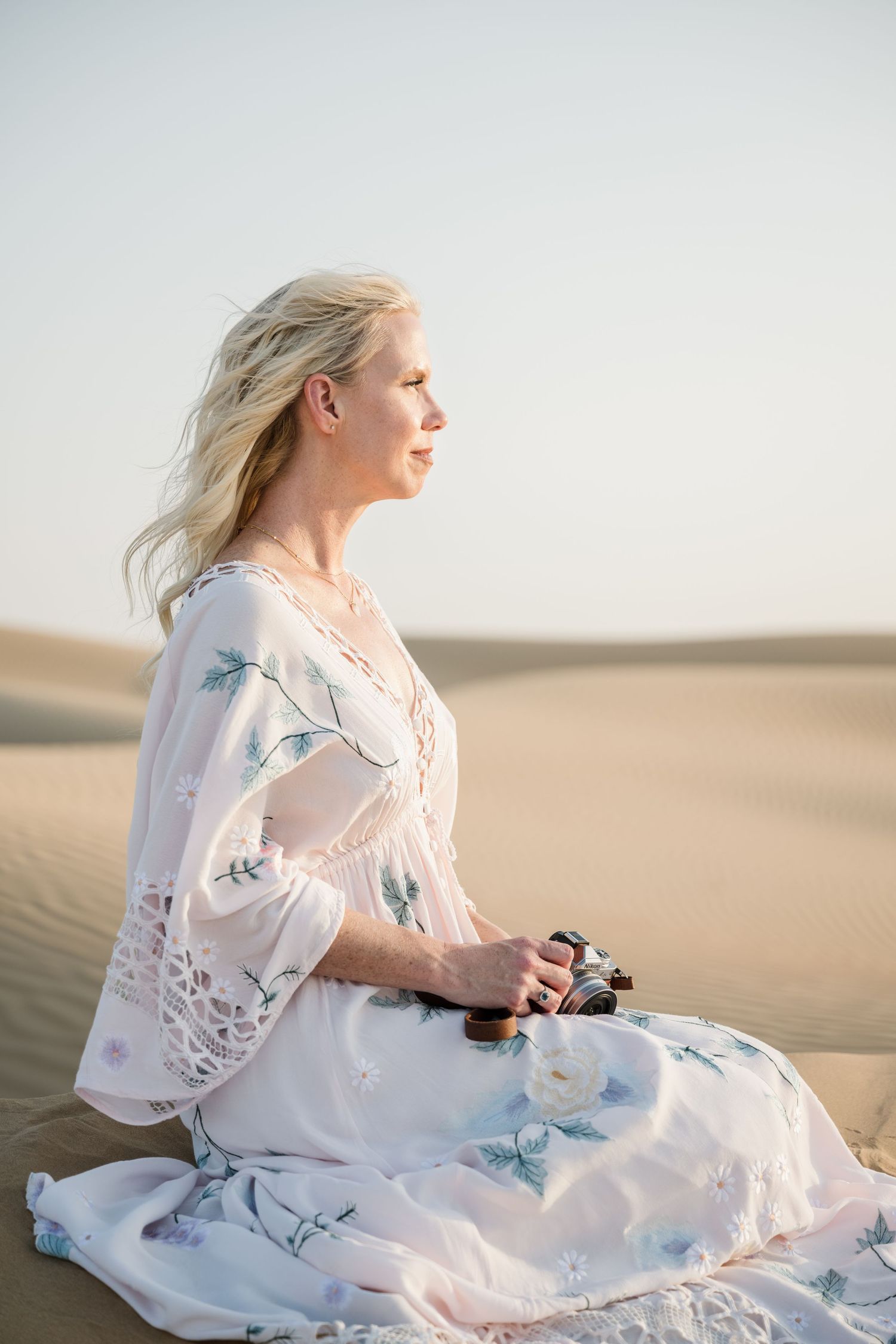 Woman in flowing white dress sitting peacefully among desert sand dunes.