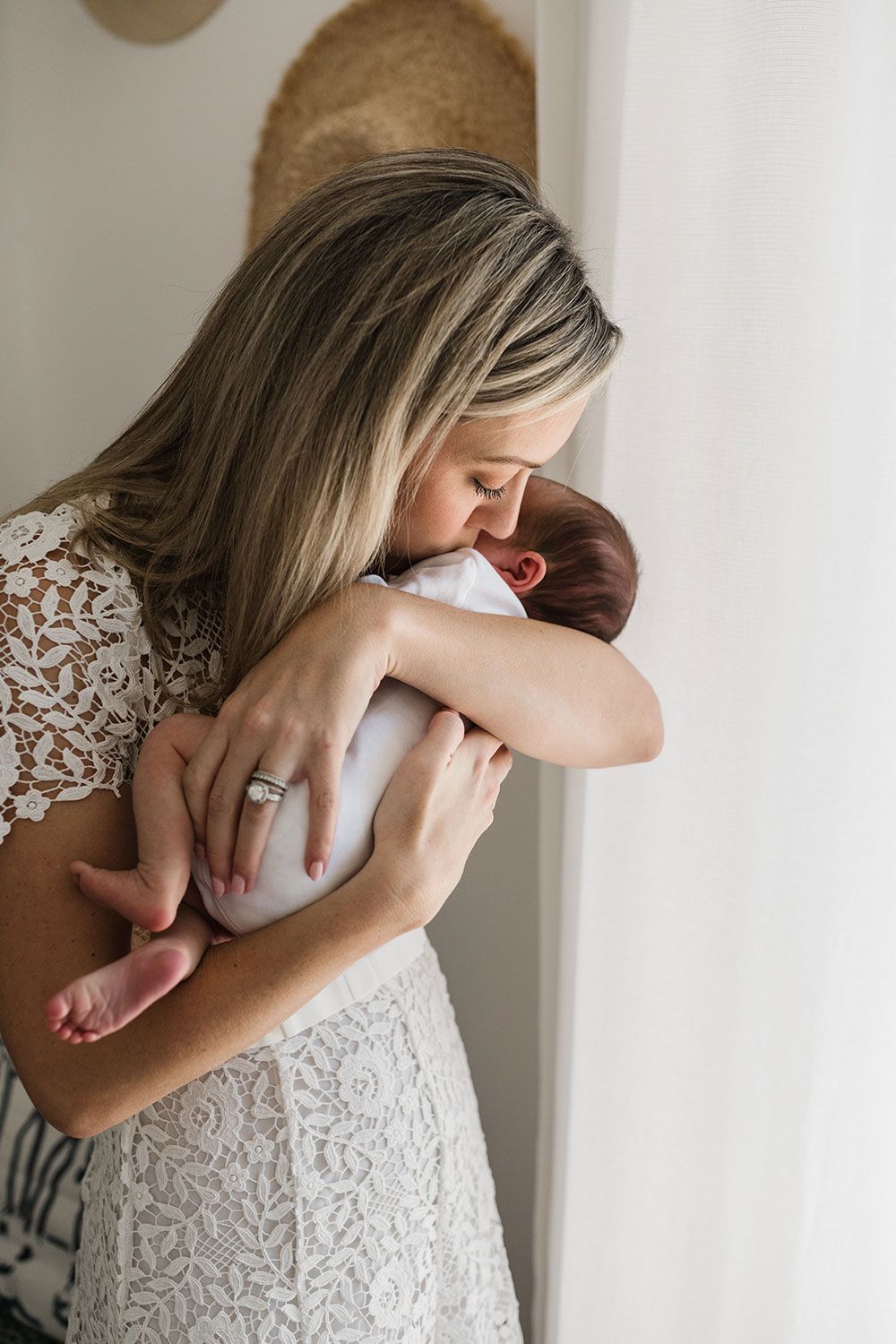 Woman in white lace dress holds newborn baby against white wall in tender embrace.