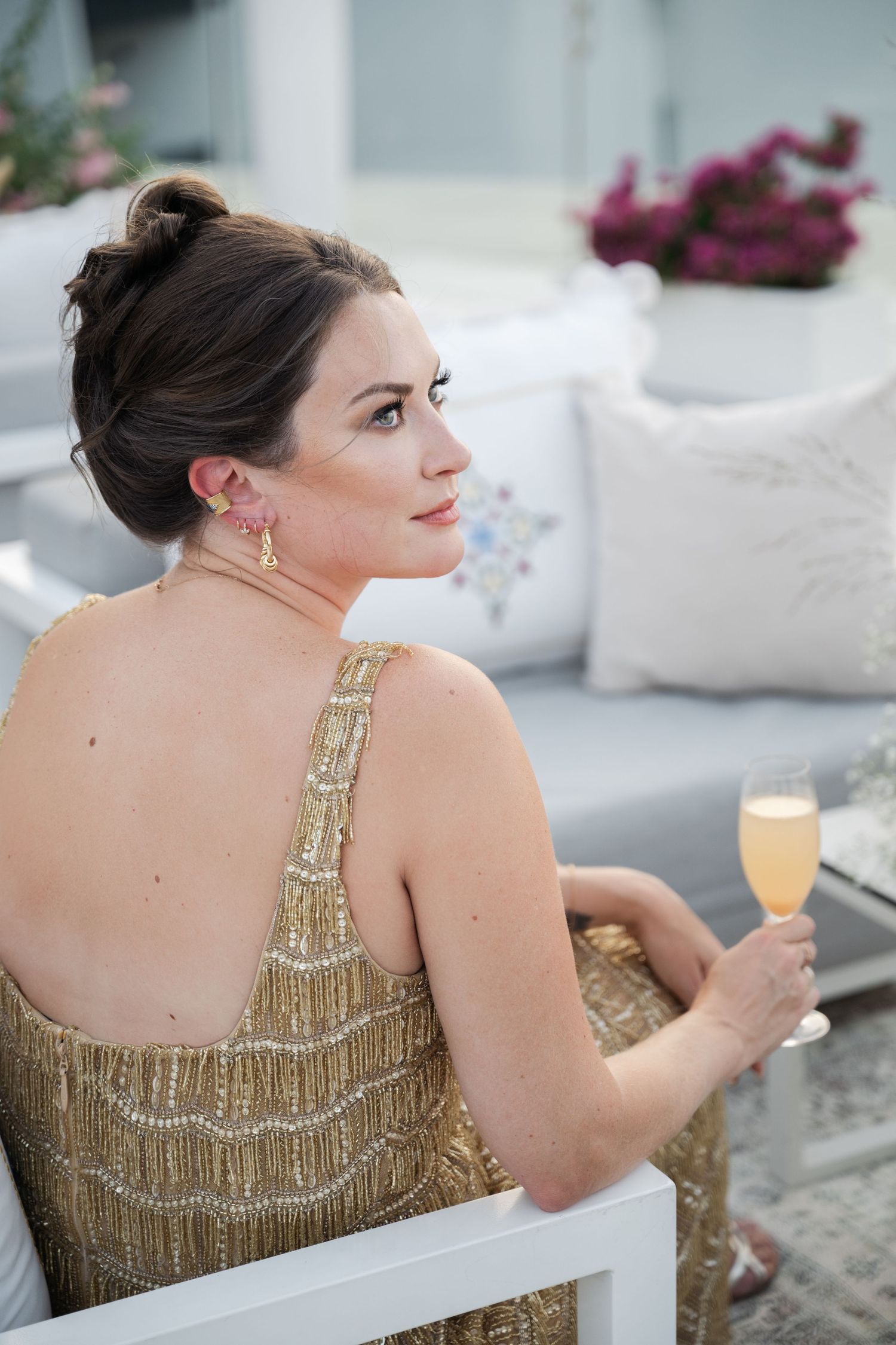 Woman in gold sequined dress relaxing at elegant outdoor venue with cocktail.