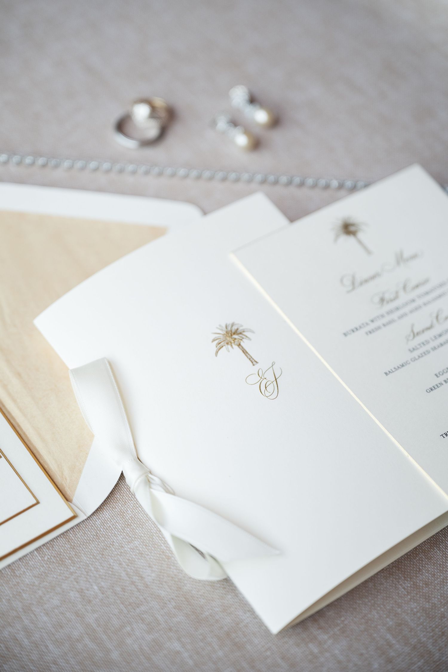 Minimalist white wedding invitations with delicate gold palm tree designs laid out on beige fabric.