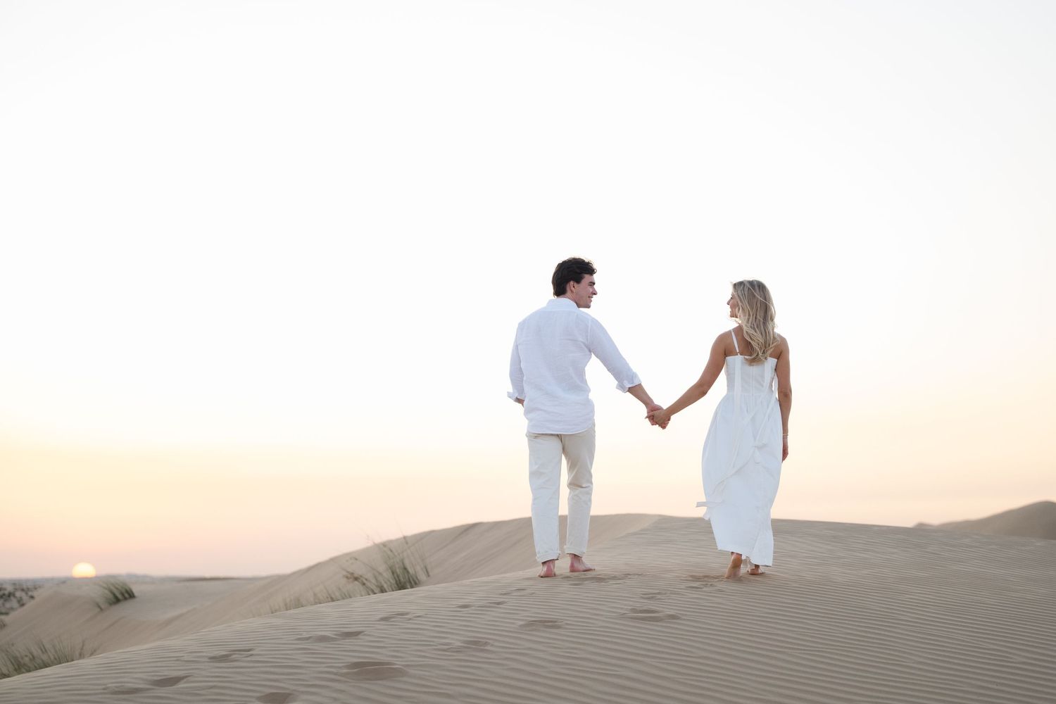 Couple in white outfits holding hands while walking on sand dunes during sunset in the desert.