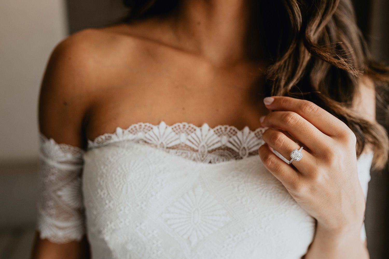 Close-up of delicate white lace wedding dress bodice with off-shoulder detail and sparkling engagement ring.