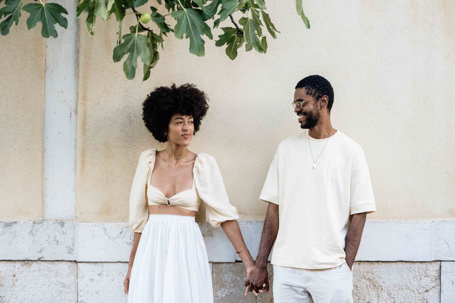 Stylish couple in white clothing stands against light wall with overhanging plants.