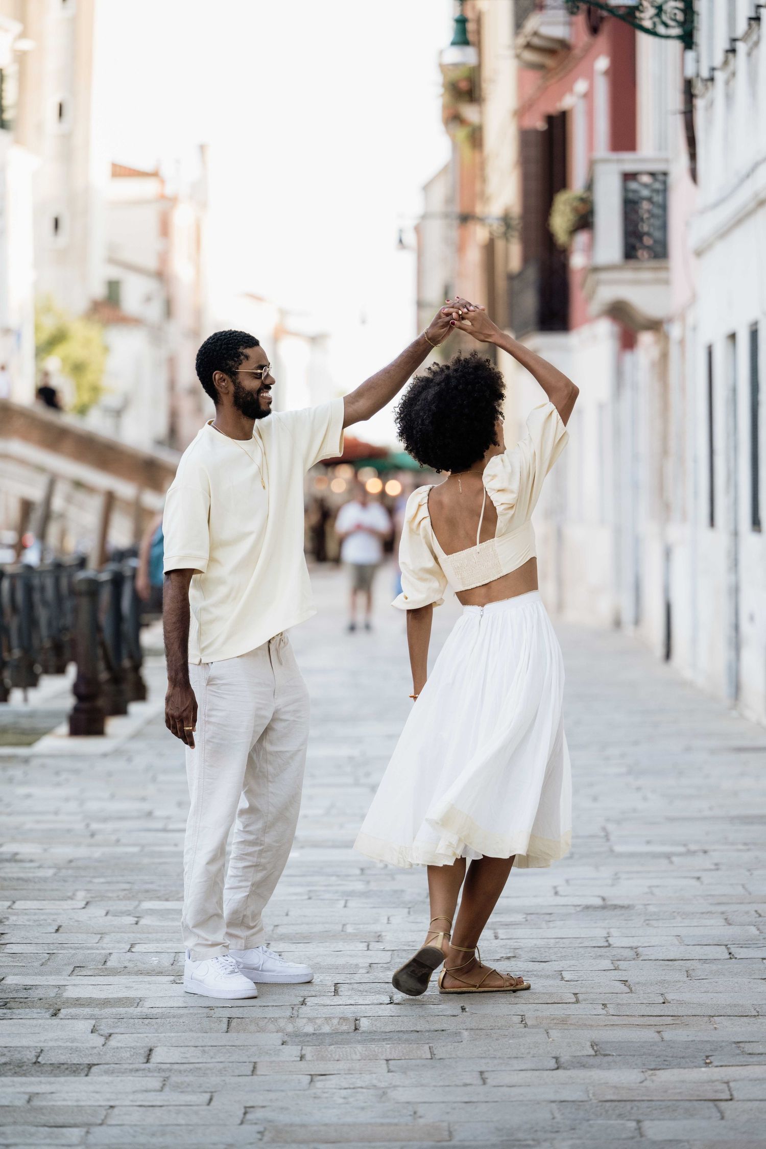 A playful couple in white outfits dancing on a historic cobblestone street.