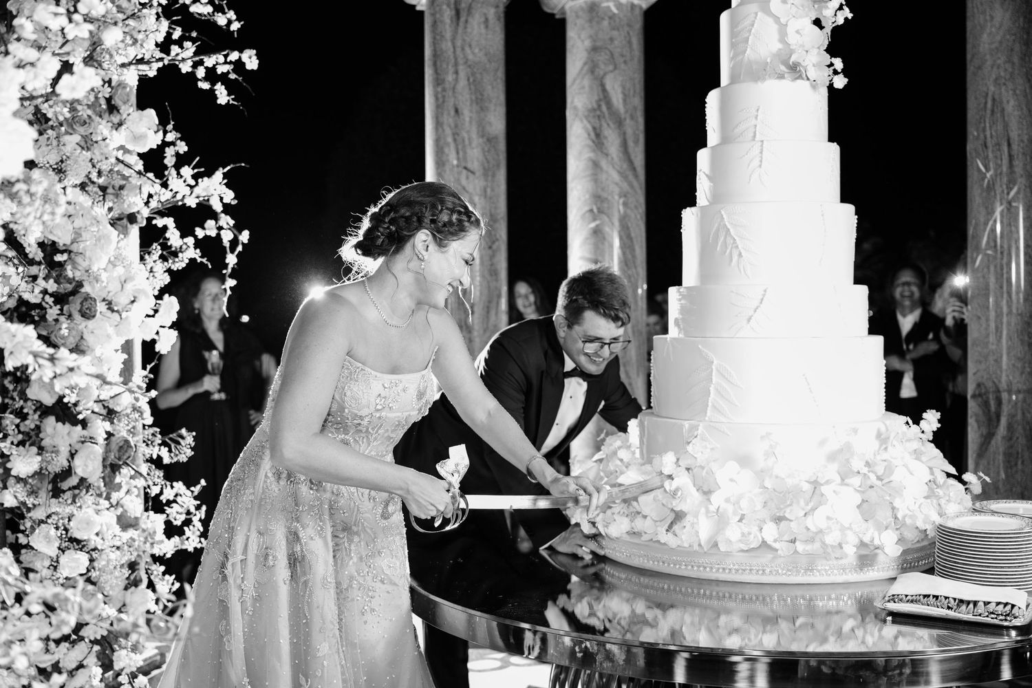 Wedding cake cutting moment captured in black and white with floral decorations in the background.